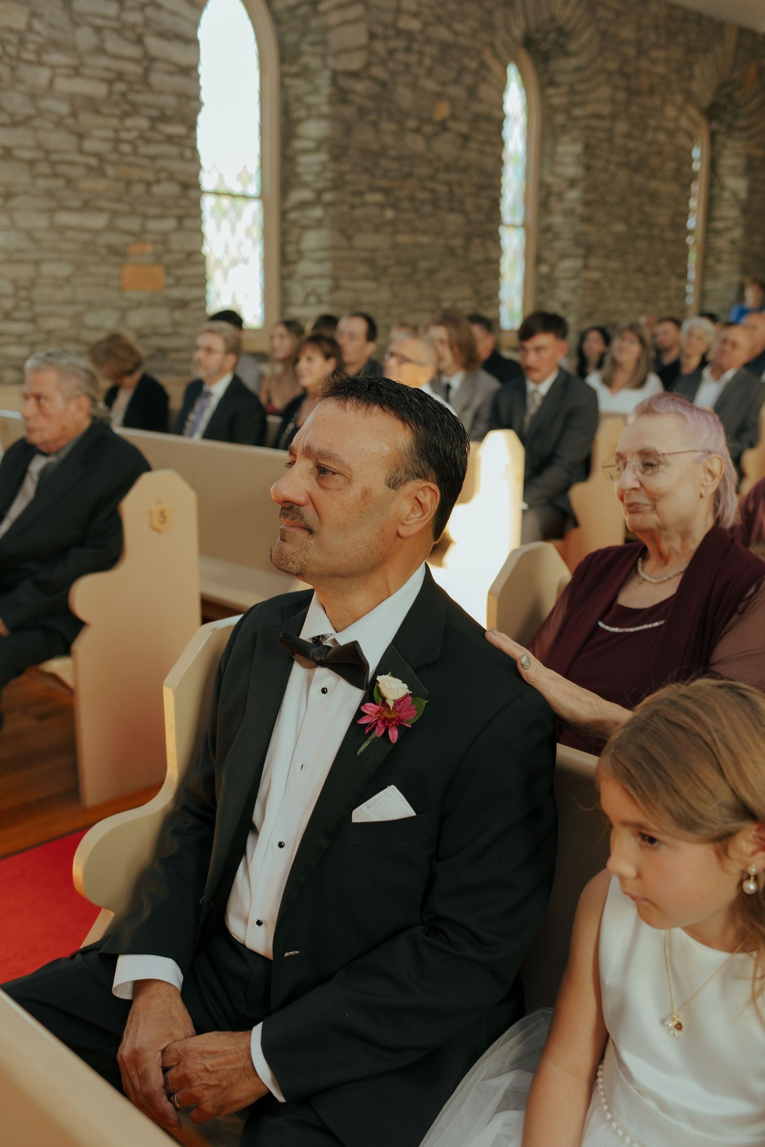 Emotional father of the bride seated in church pew during wedding ceremony in stone chapel.