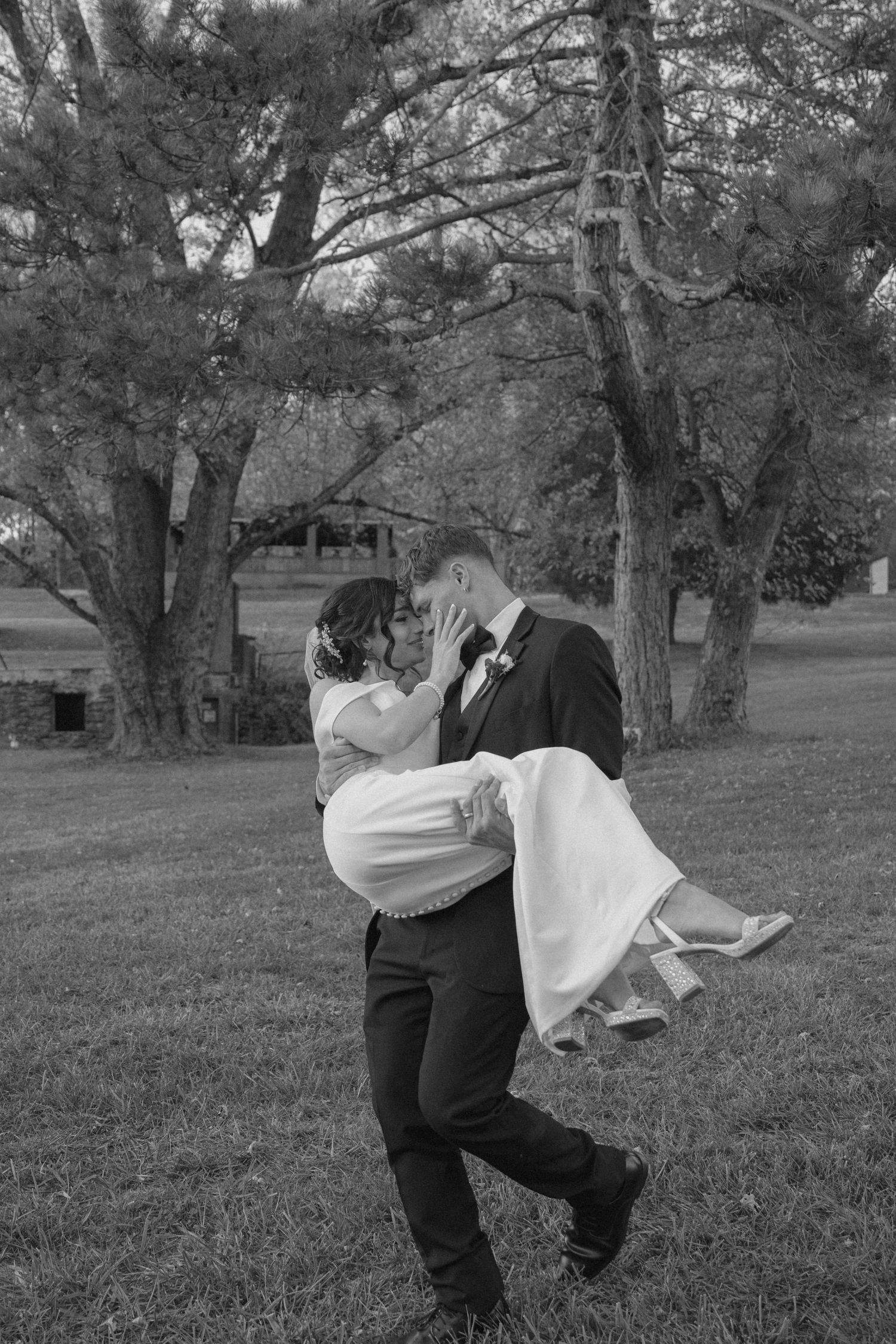 Black and white photo of groom lifting bride in wedding dress outdoors beneath tall trees.
