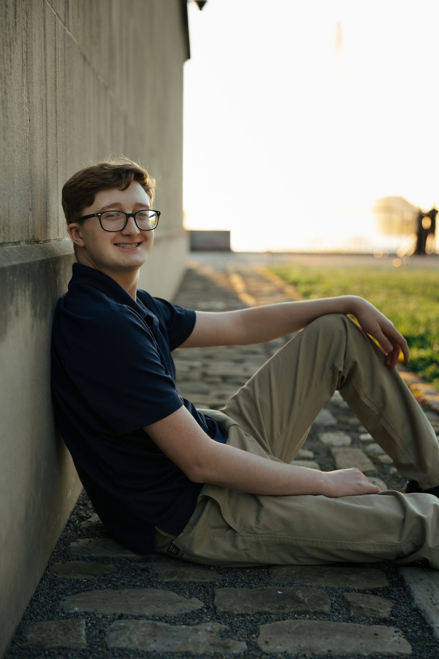 Smiling teen boy with glasses sitting relaxed against a wall on stone pavement at golden hour.