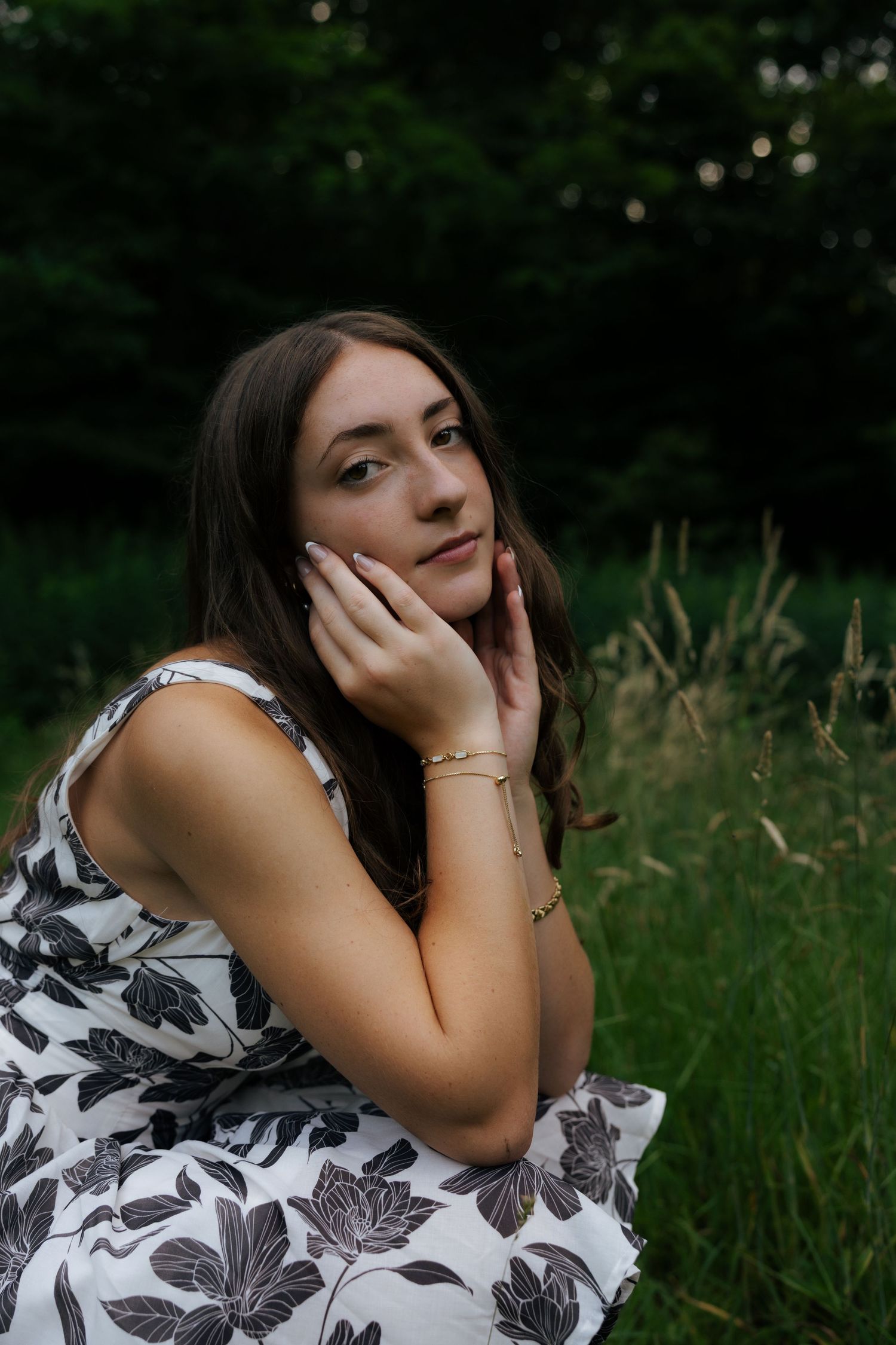 Young woman in floral dress posing with hands near face wearing gold bracelet in nature.