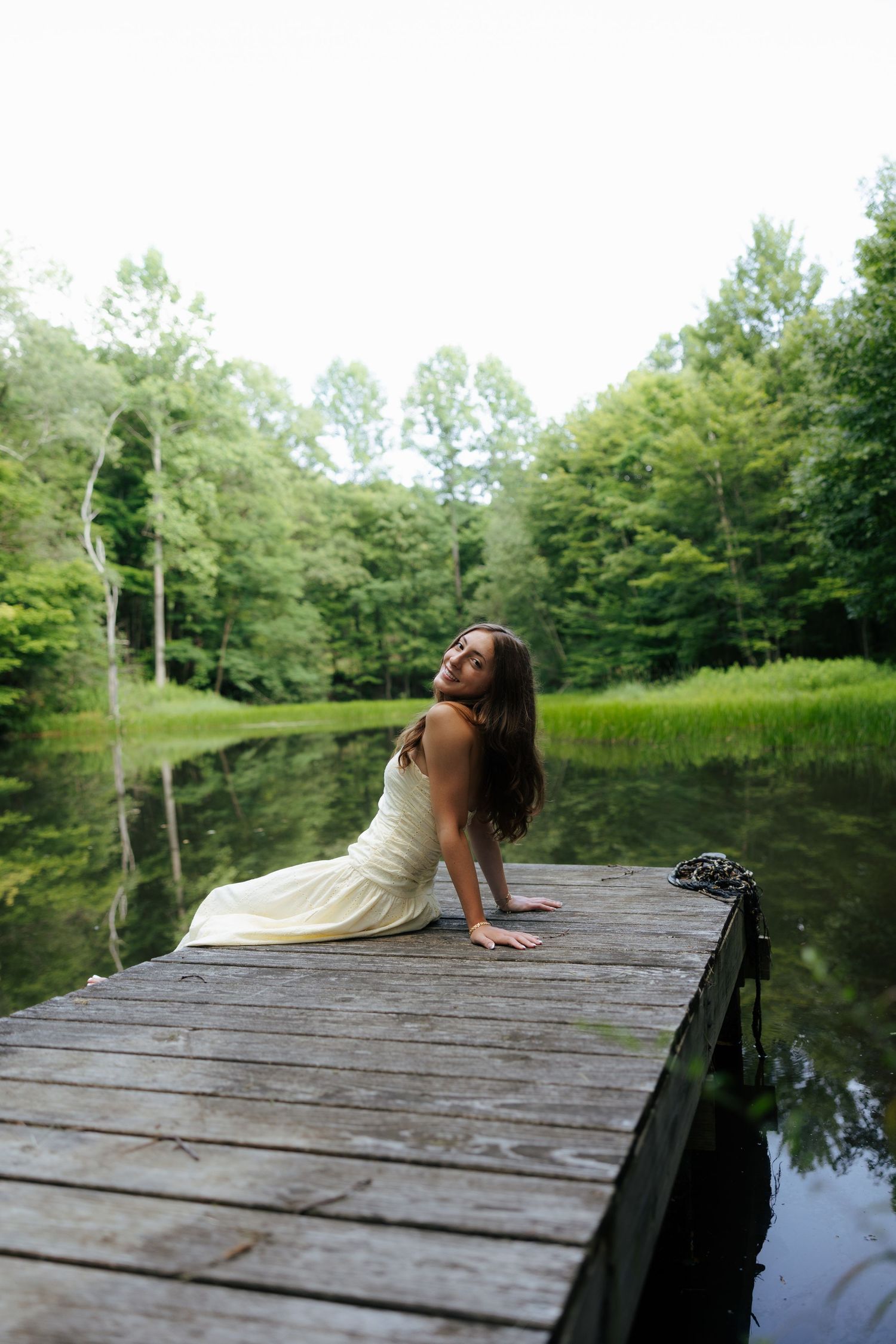 Woman in white dress sitting on wooden dock by serene forest lake.