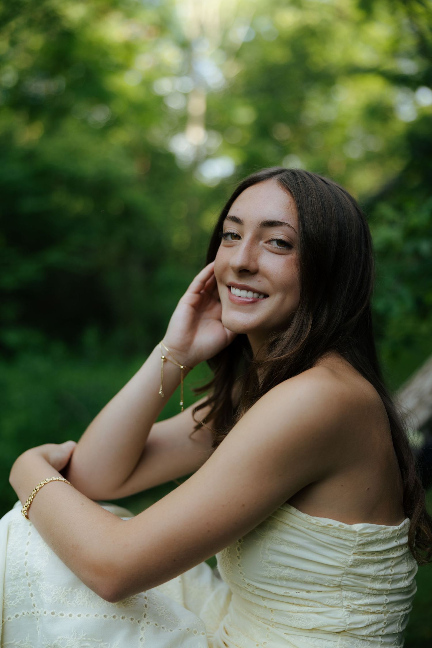 Smiling young woman in cream strapless dress with gold bracelets posing outdoors in green forest.