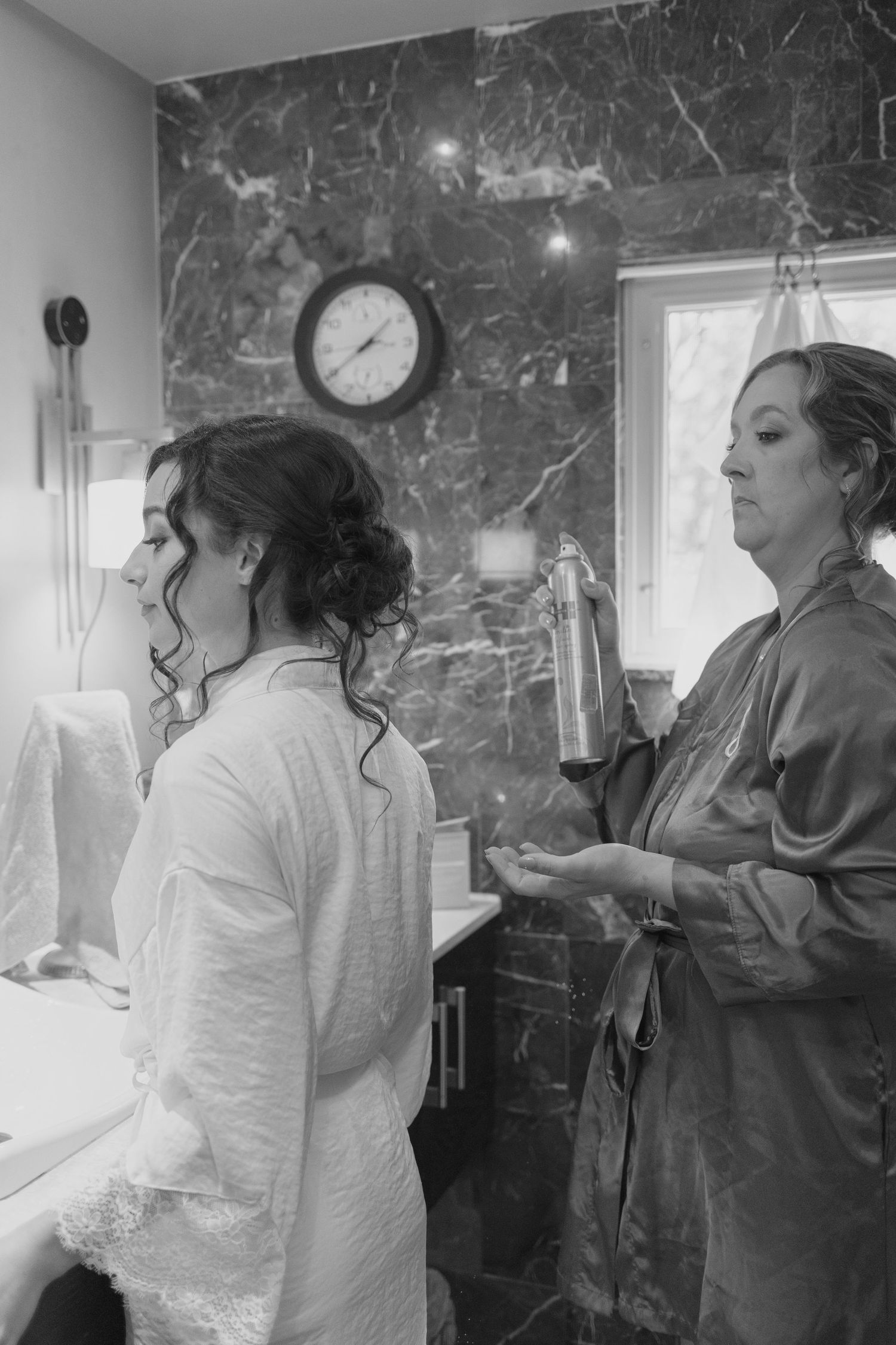 Black and white photo of bride in robe getting hair styled in marble bathroom before wedding.