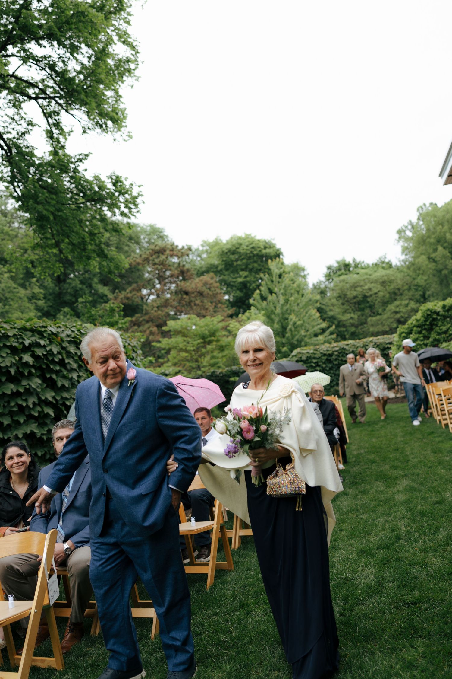 Elderly couple walks down wedding aisle outdoors, woman holding pink and white floral bouquet.