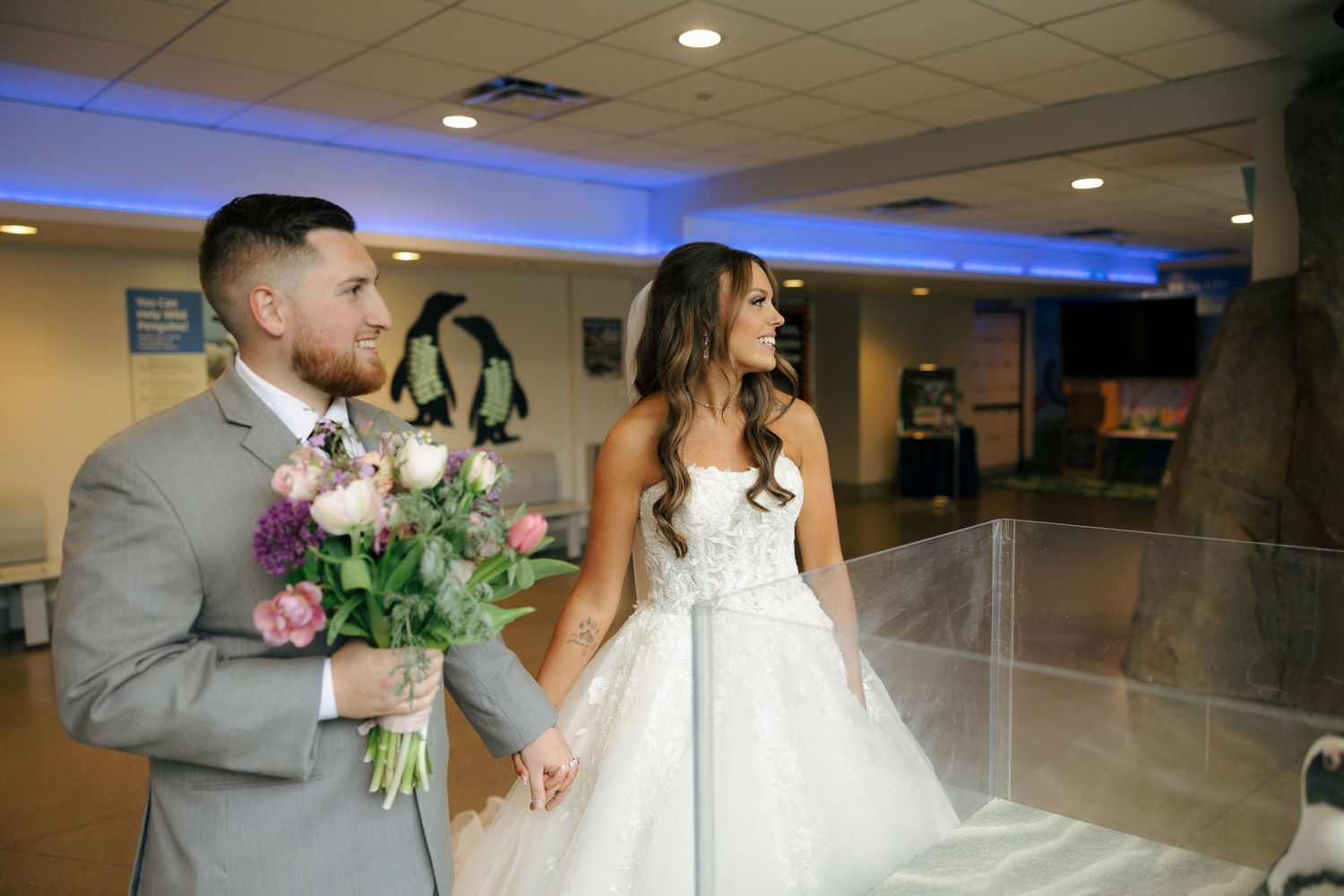 Bride in white lace gown and groom in gray suit holding pink and purple bouquet at indoor wedding venue.