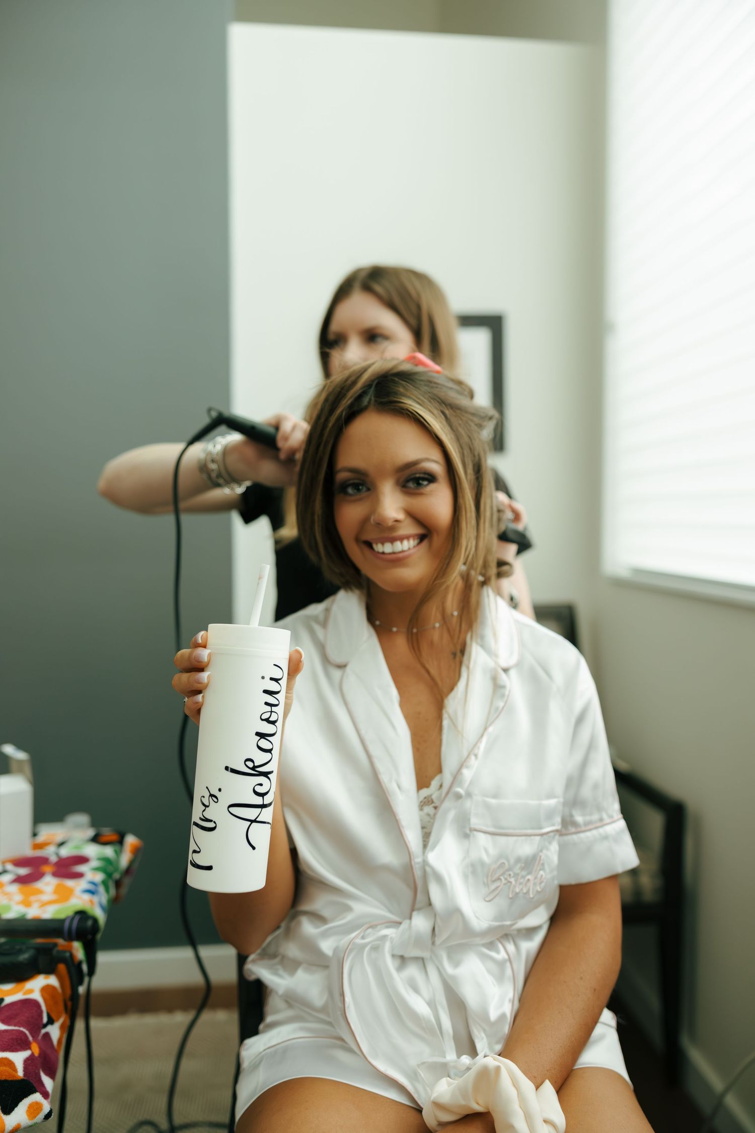 Smiling bride in white satin robe holds personalized cup while getting hair styled by a hairdresser.