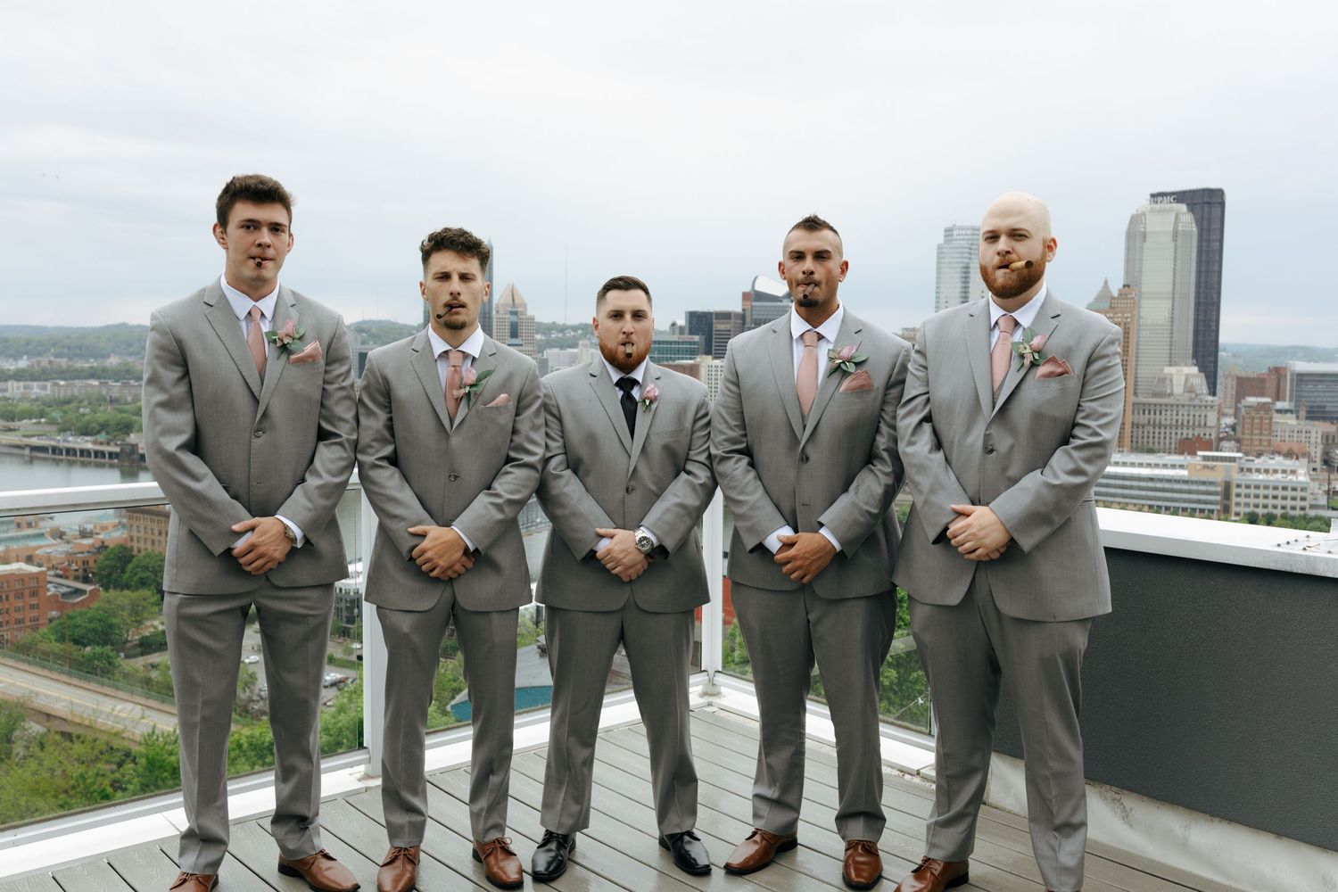 Five groomsmen in gray suits with floral boutonnieres pose on a rooftop with city skyline backdrop.
