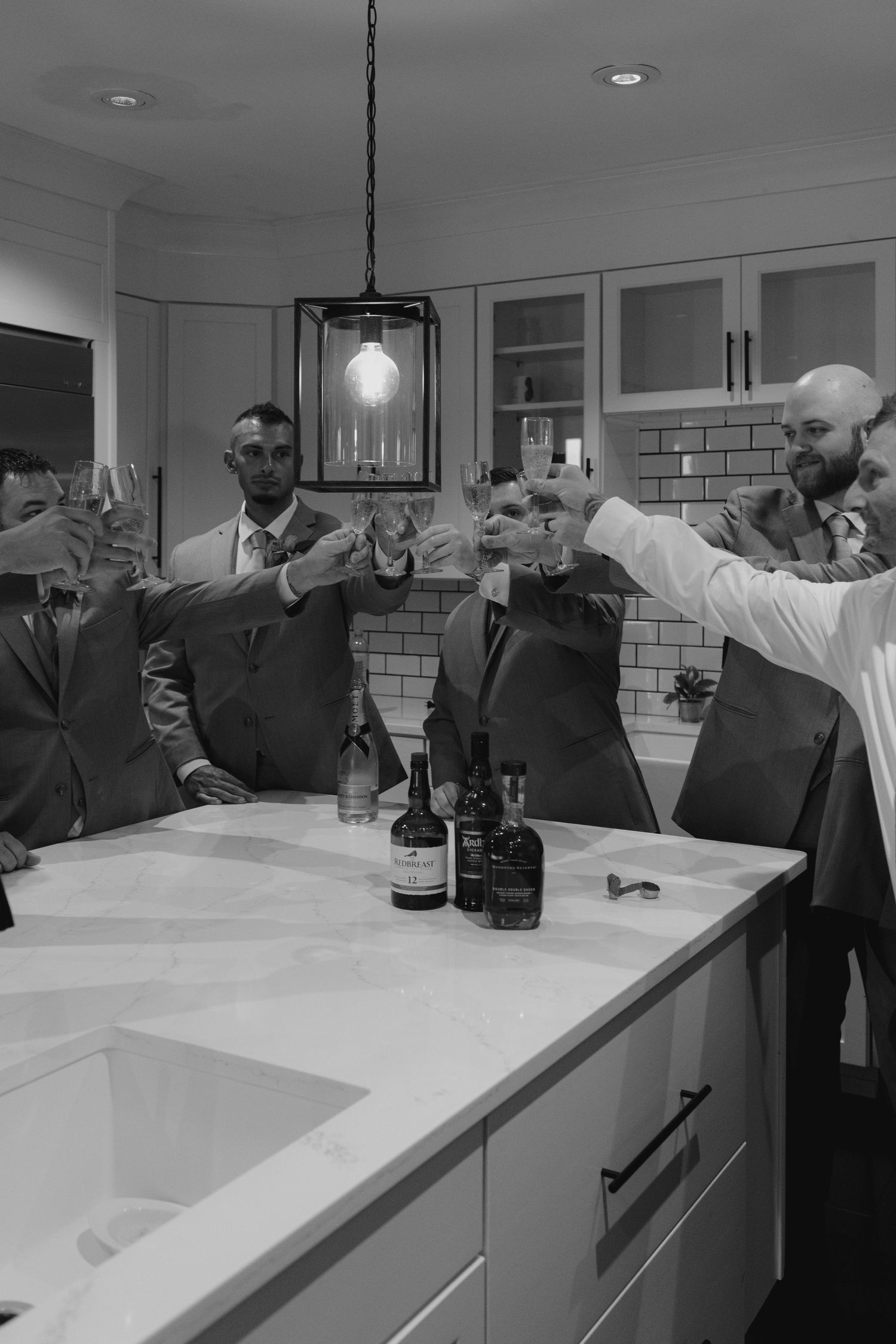 Black and white photo of groomsmen raising glasses in a toast around a kitchen island with whiskey bottles.
