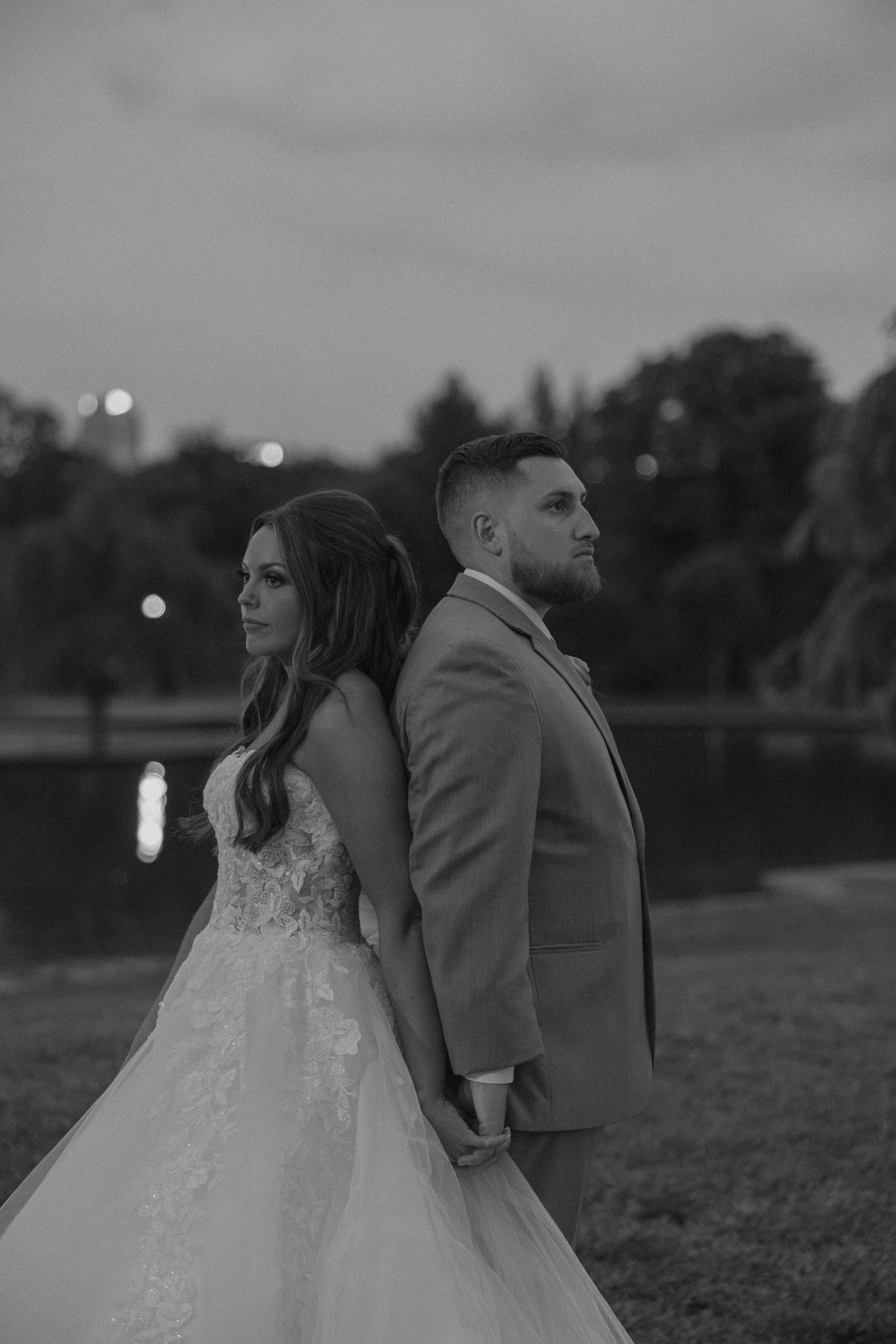 Black and white portrait of bride and groom standing back to back at twilight near a serene lake.
