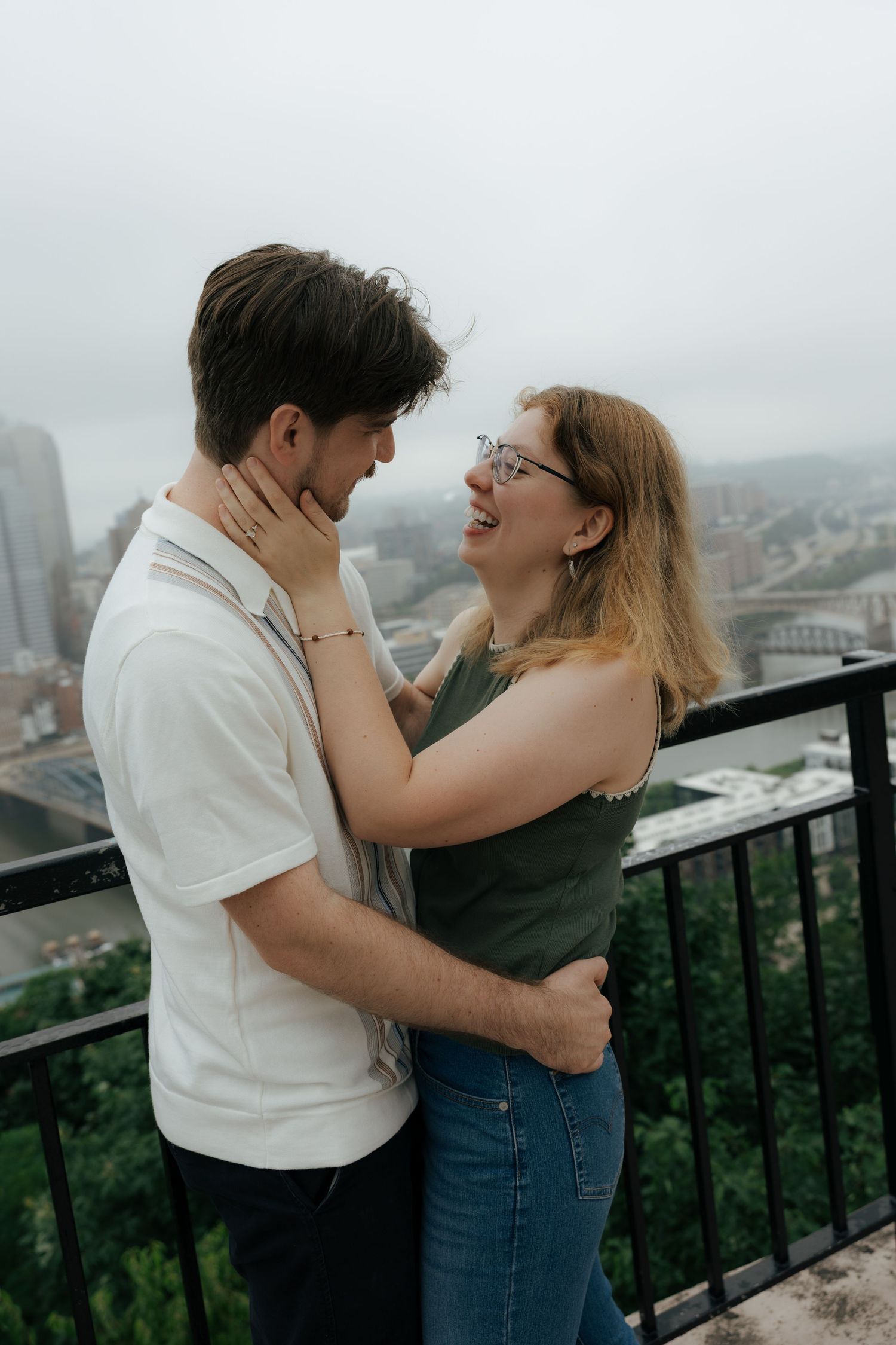 Couple embracing on a rooftop overlook, woman laughing joyfully while holding mans face, city skyline in background.