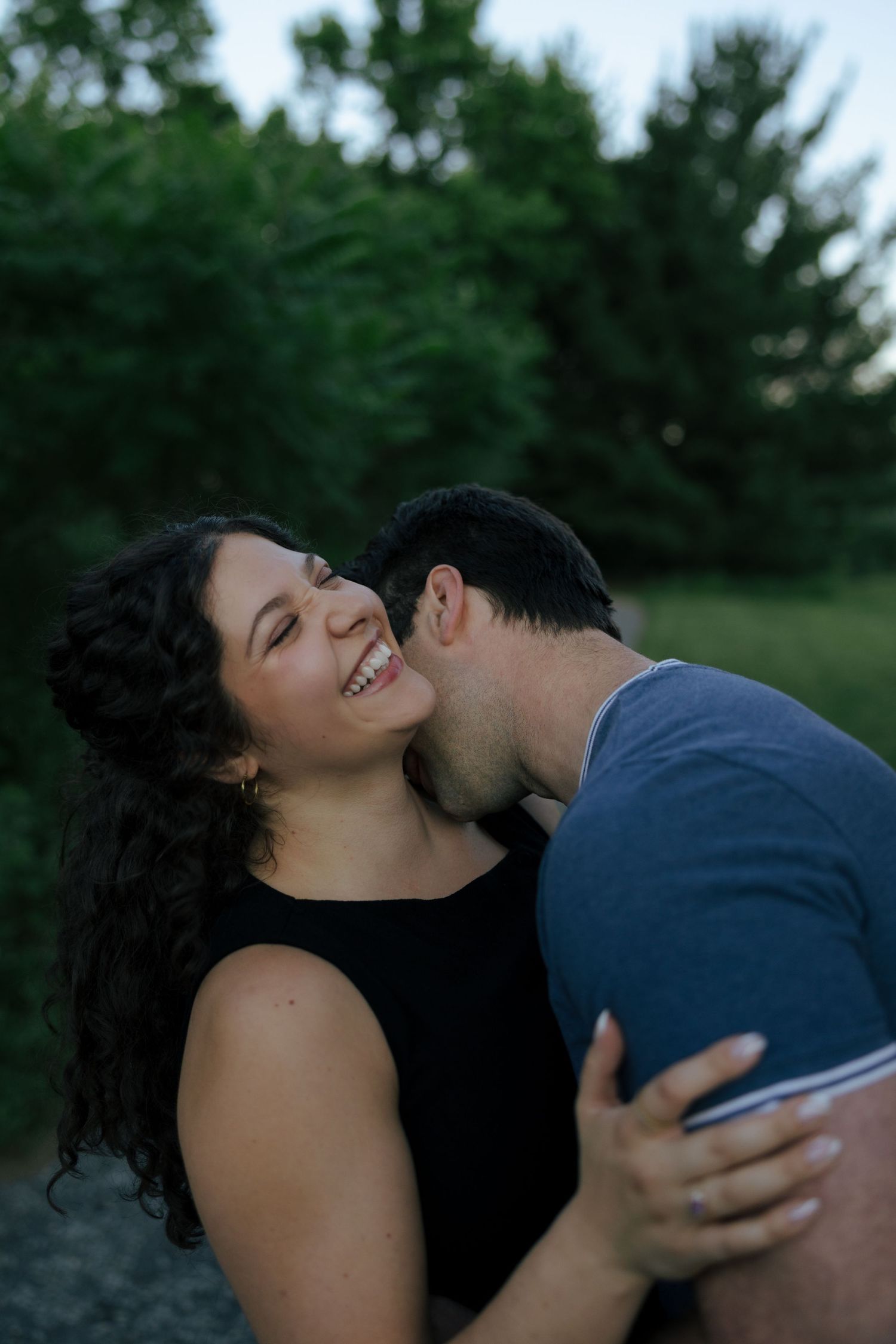 A happy woman smiles as her partner kisses her cheek while embracing outdoors with green trees in the background.