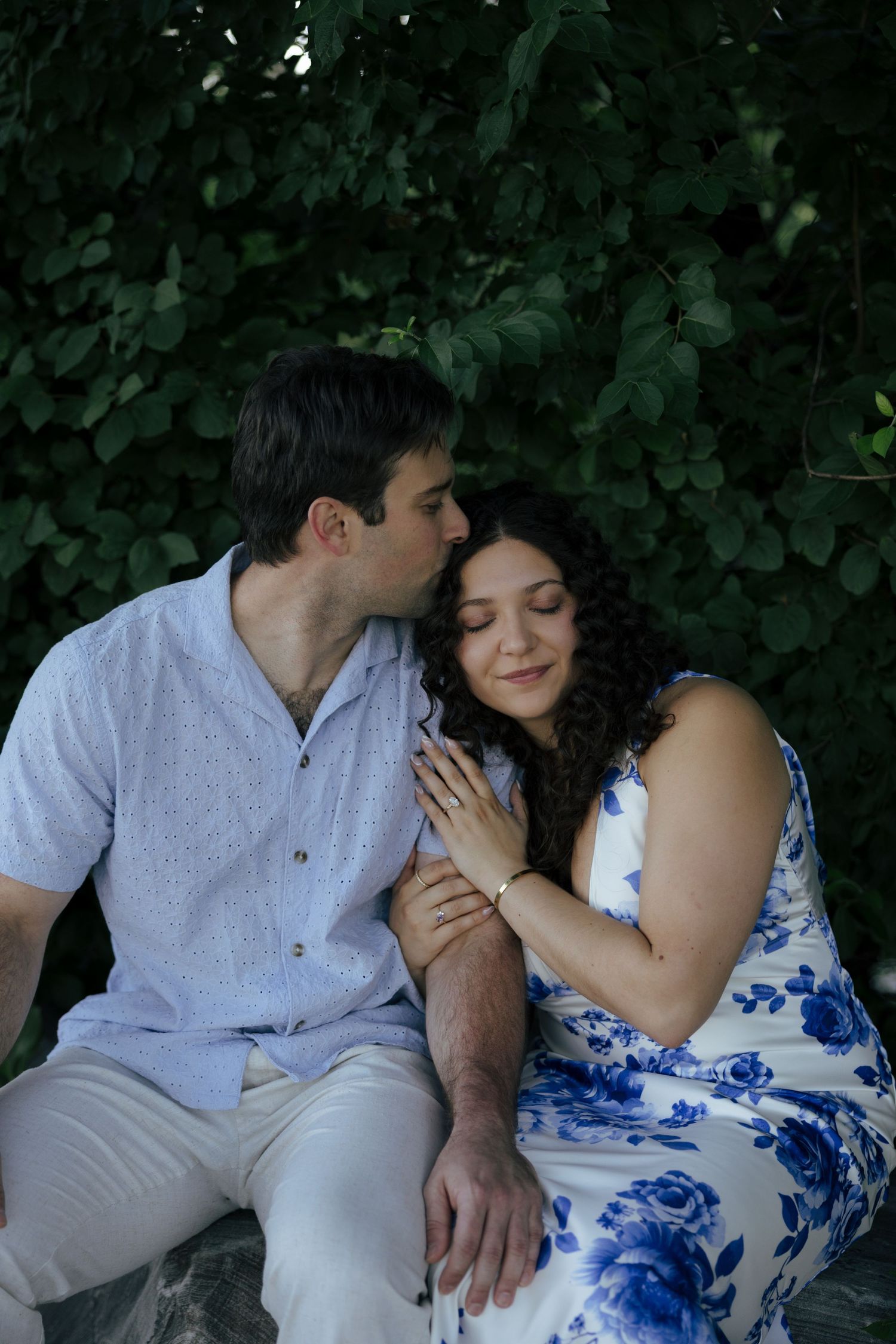 Couple sitting on rocks surrounded by lush greenery, man kissing woman on cheek, woman in blue floral dress smiling softly.