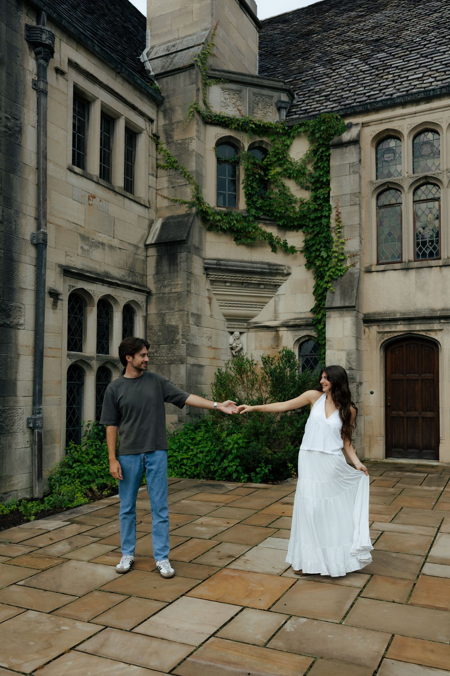 Romantic couple holding hands in historic stone castle courtyard with ivy-covered gothic architecture.