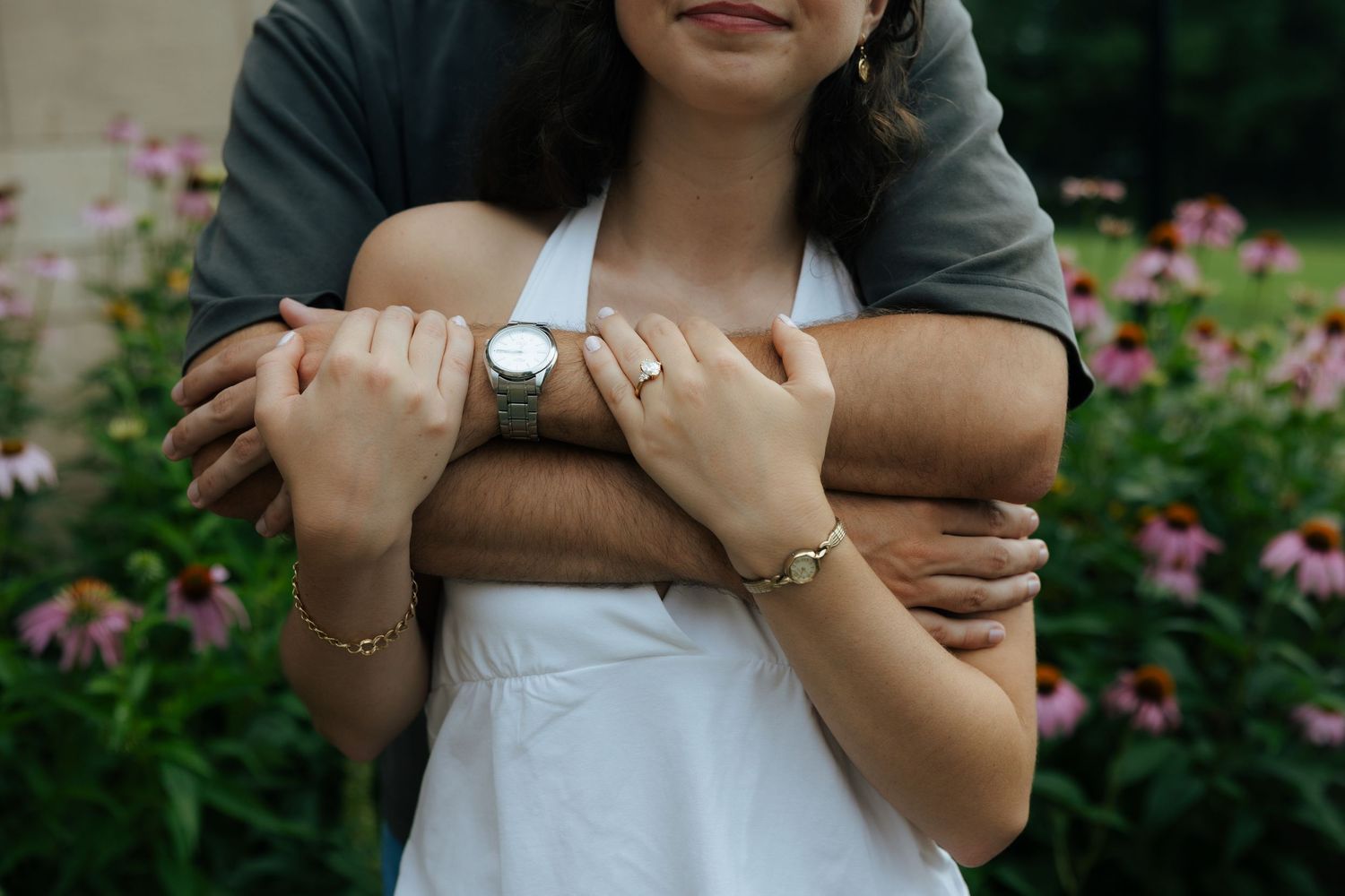 A man wraps his arms around a woman in a white dress, hands clasped, in front of pink garden flowers.