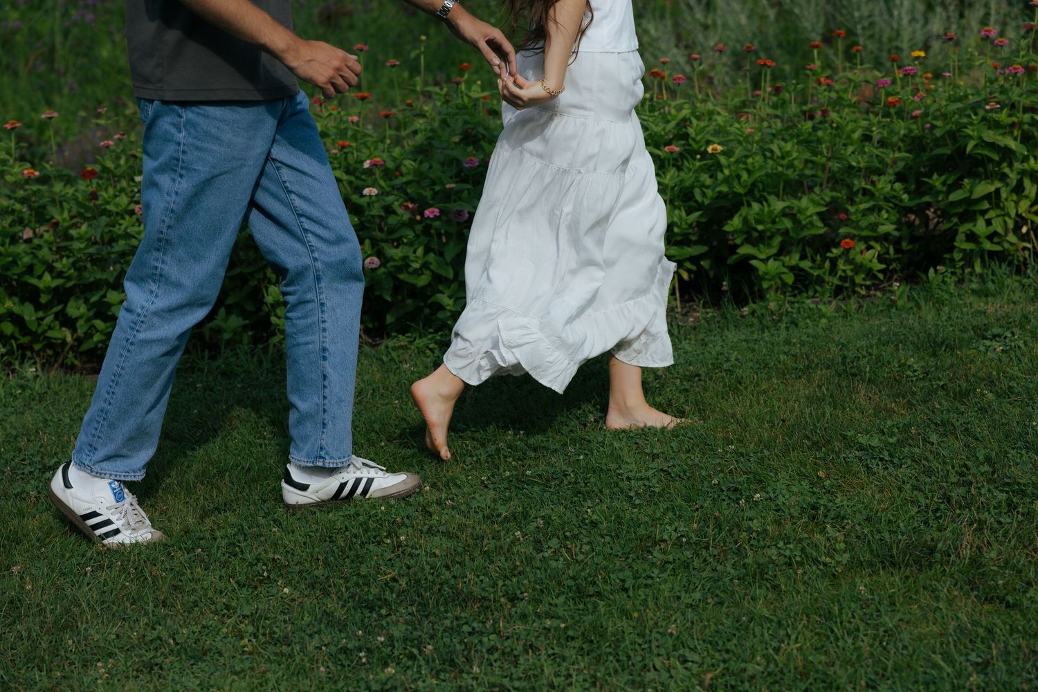 Couple walking barefoot on green grass, woman in flowing white dress holding mans hand.