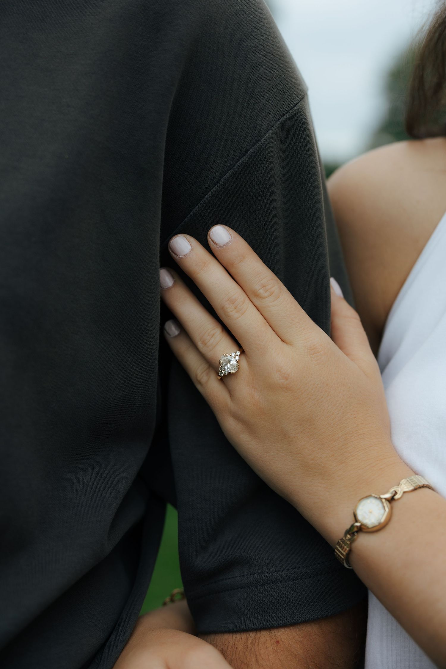 Close-up of woman's hand with diamond engagement ring and gold bracelet resting on man's gray shirt.