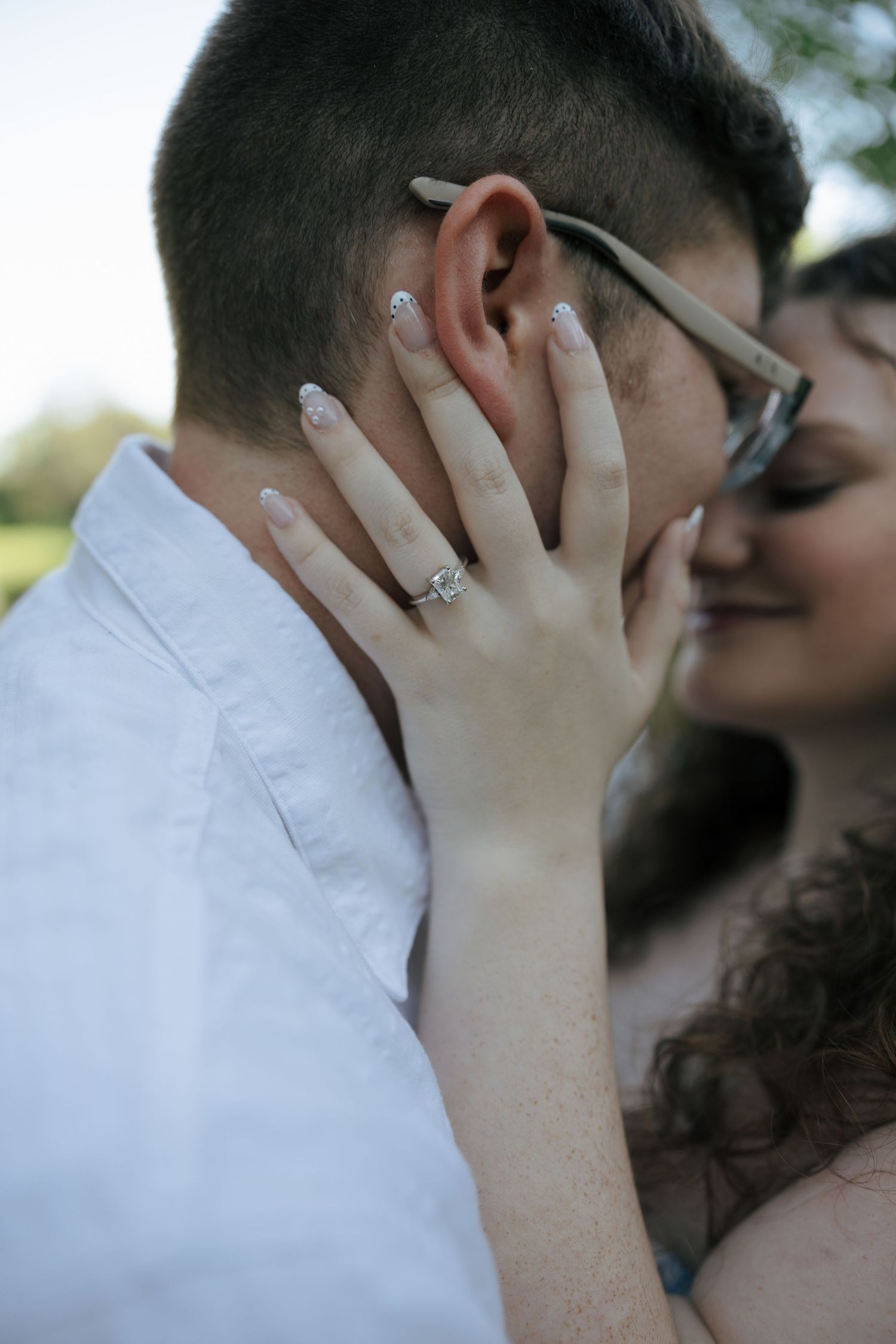 Close-up of womans hand showing diamond engagement ring while tenderly touching her partners face near his glasses.
