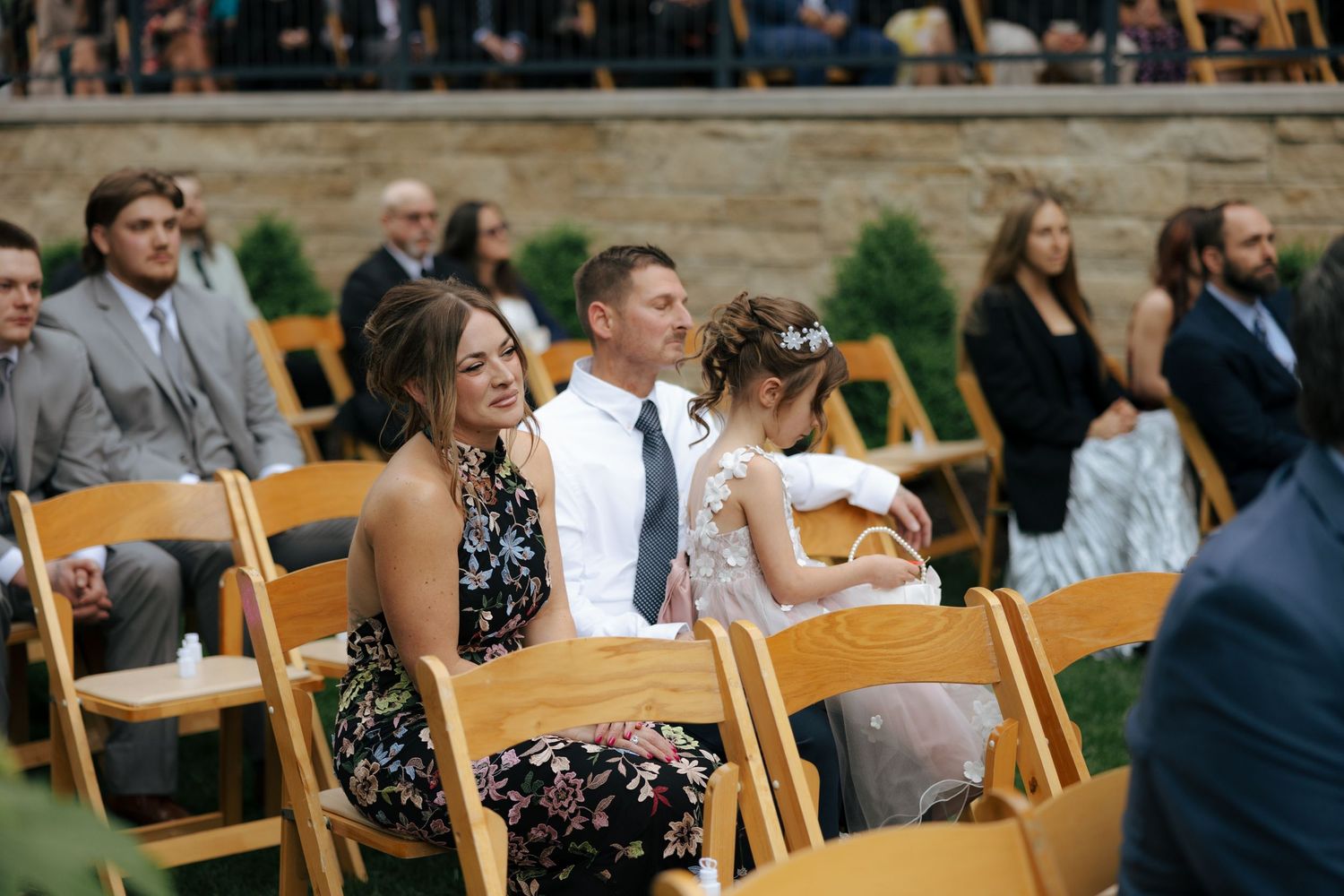 Wedding guests seated in wooden chairs smile during an outdoor ceremony.