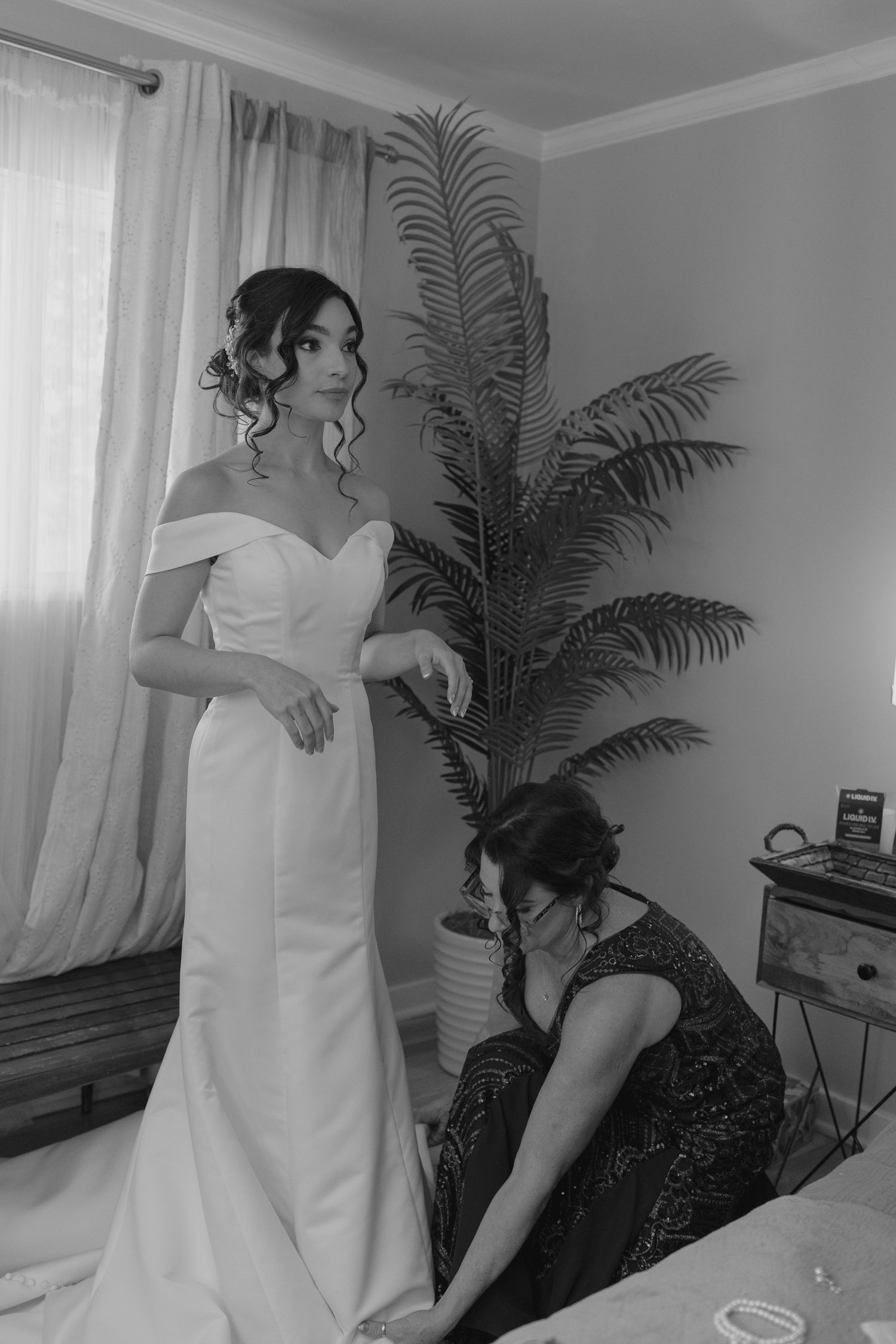 Bride in off-shoulder white gown being helped by bridesmaid near a window in black and white.
