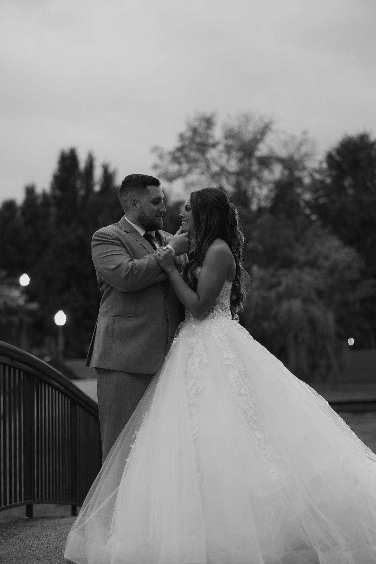 Black and white portrait of bride and groom sharing a romantic moment on a bridge at twilight.