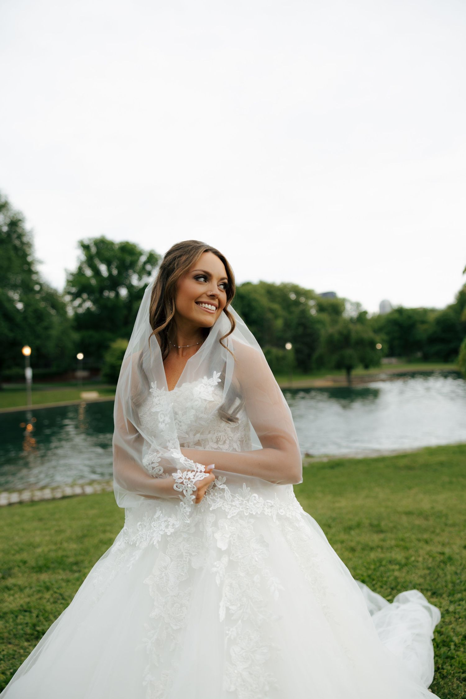 Smiling bride in lace wedding gown and veil poses outdoors near a pond surrounded by lush green trees.