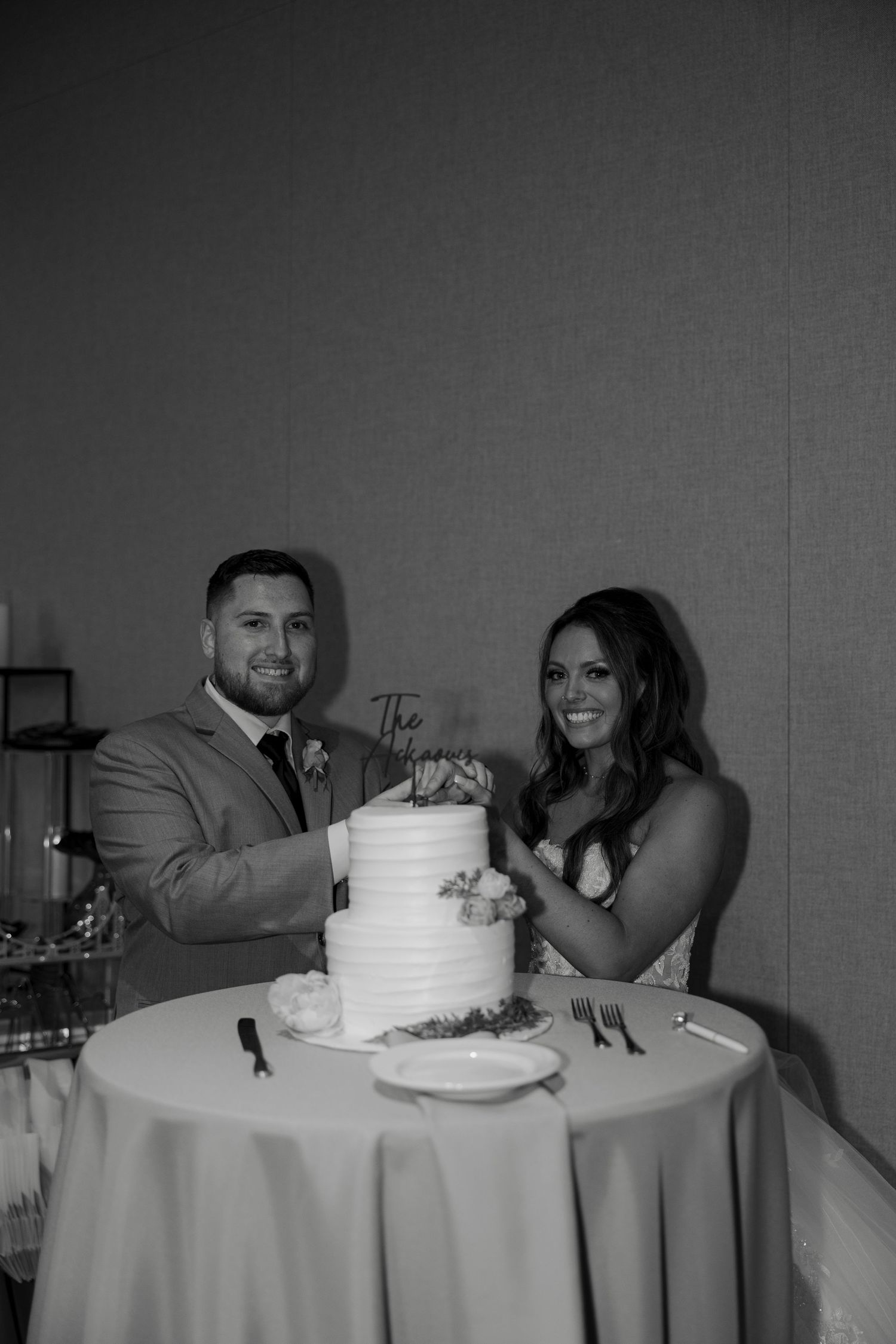 Bride and groom smiling while cutting their white tiered wedding cake at reception table in black and white photo.