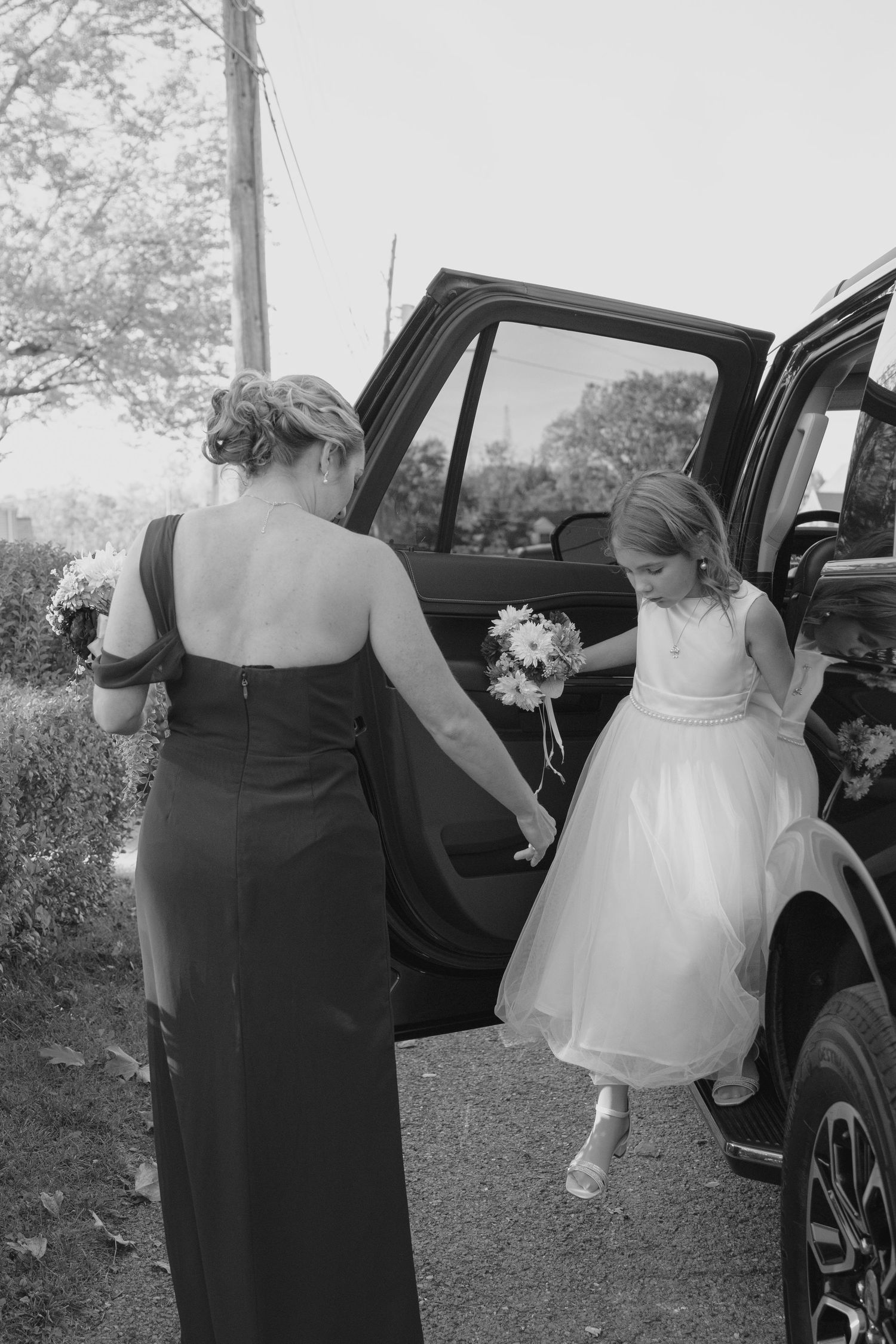 Black and white photo of bridesmaid helping young flower girl in white dress exit black SUV before wedding ceremony.