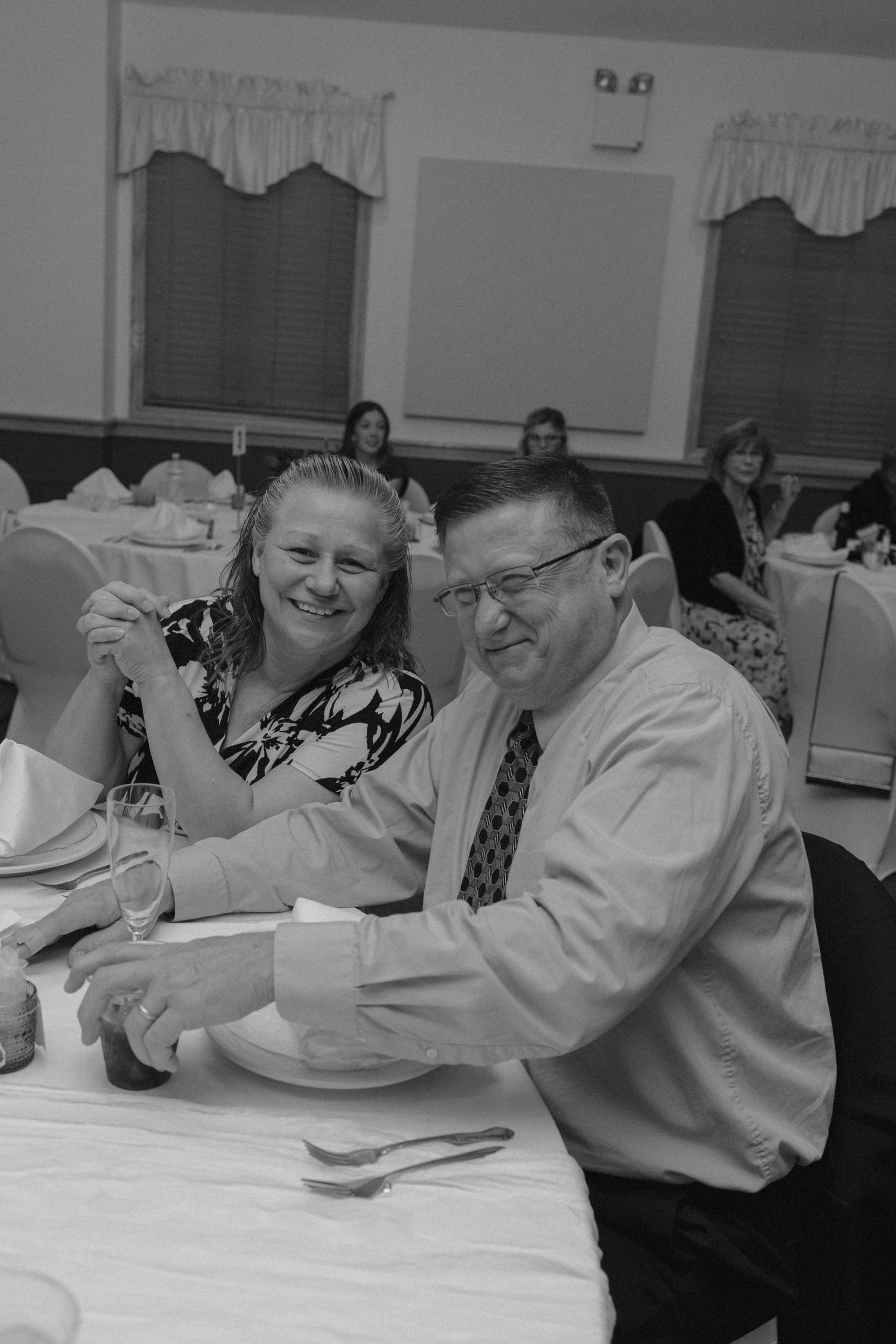 Smiling couple enjoys a cheerful moment together at a wedding reception dinner table in black and white.