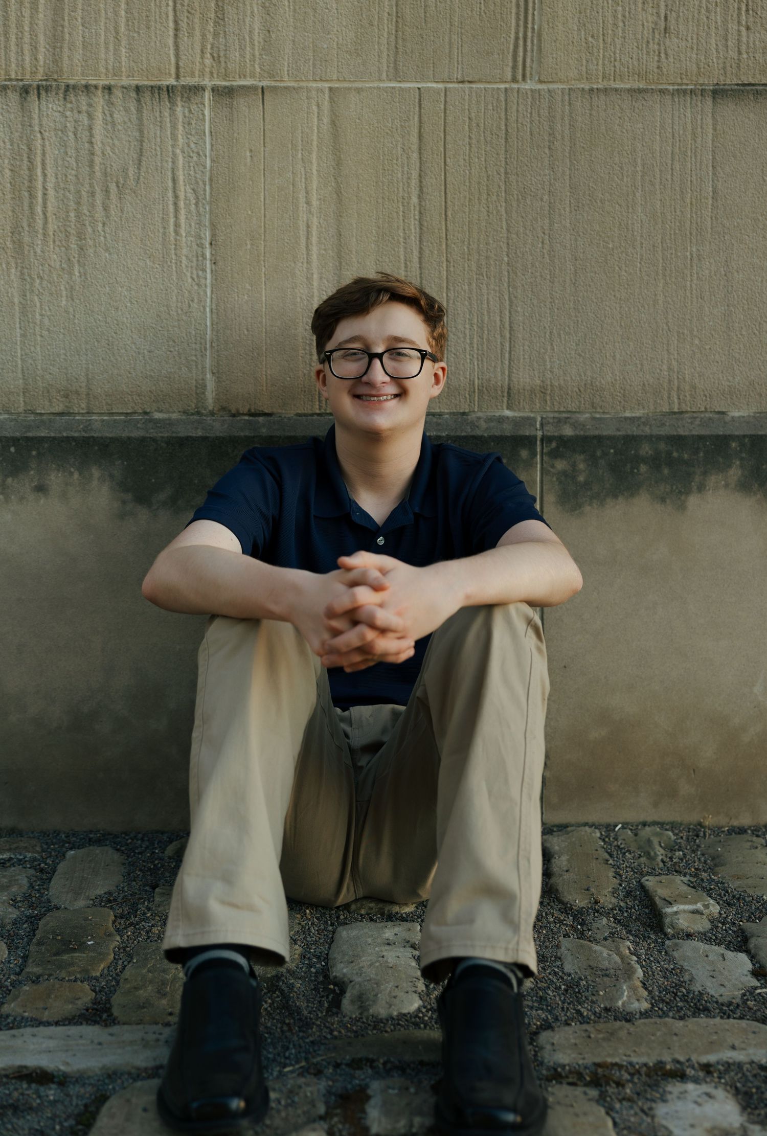 Cheerful teen boy with glasses and braces sits cross-legged on cobblestone steps against a wall.