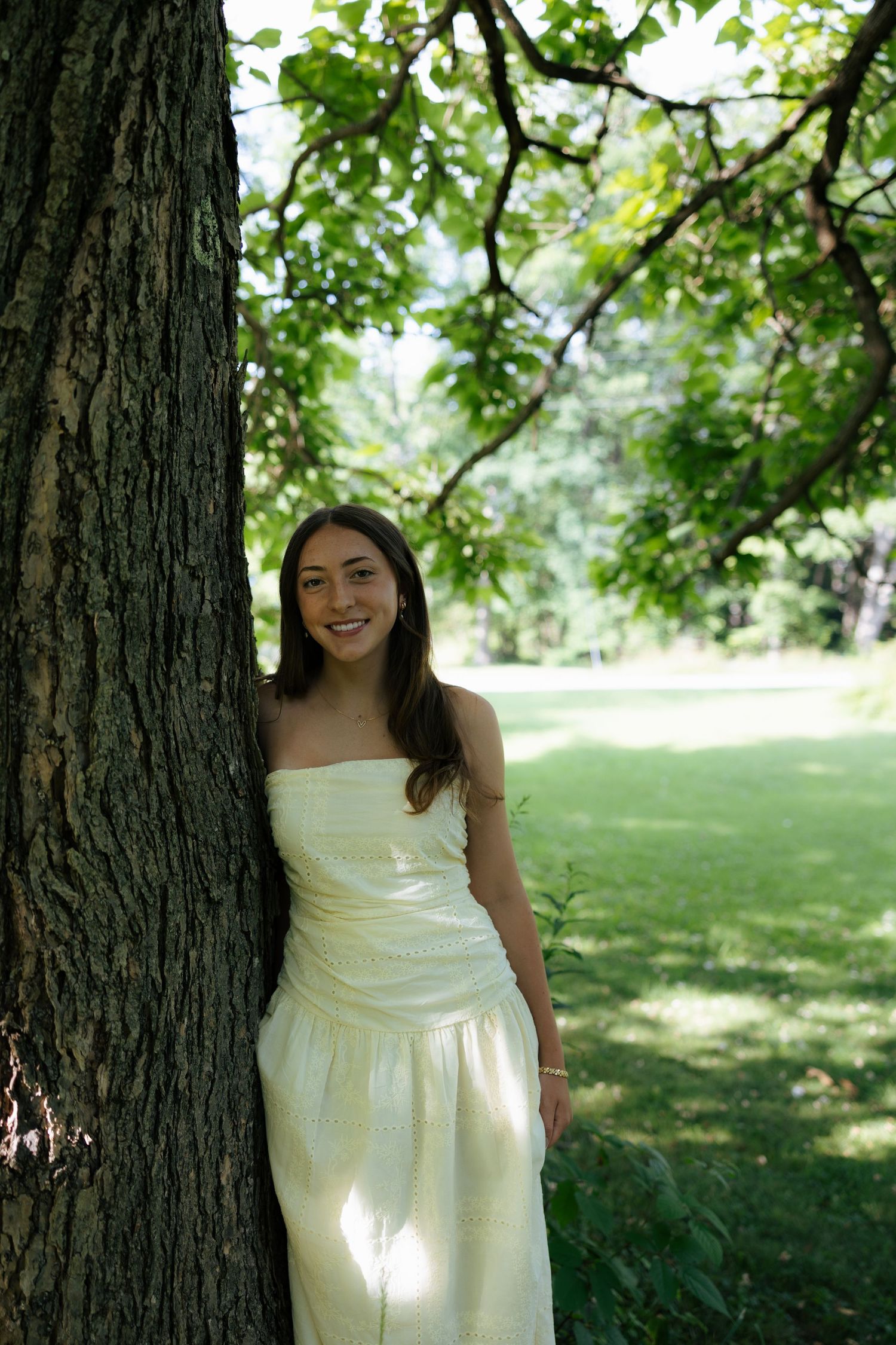 Smiling young woman in cream strapless dress leaning against large tree trunk in sunny park.