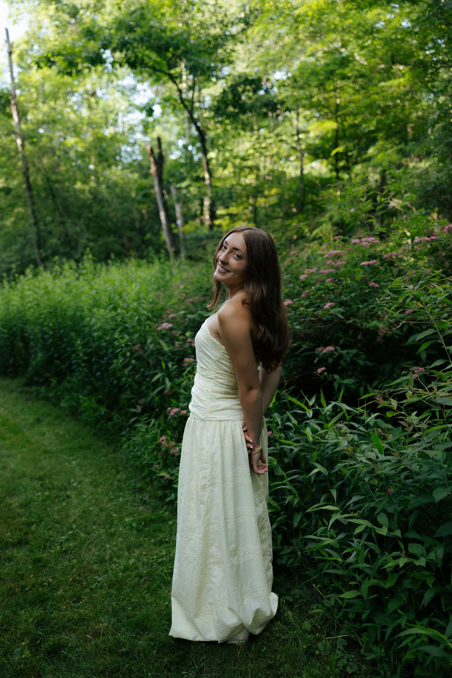 Young woman in flowing white strapless dress standing in lush green garden among wildflowers.