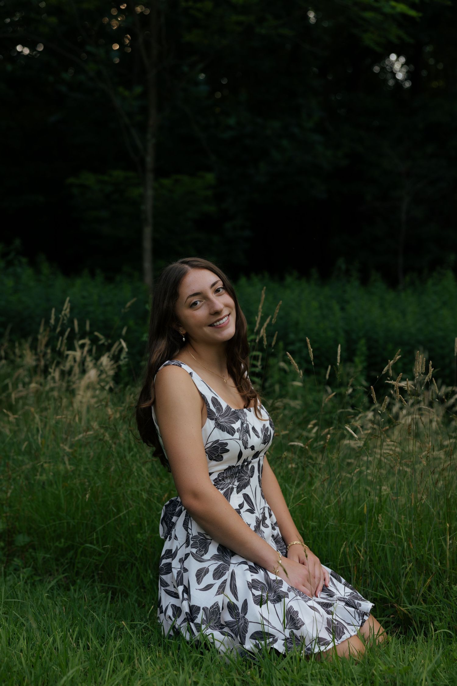 Young woman in floral dress sitting gracefully on green grass in front of lush forest backdrop.