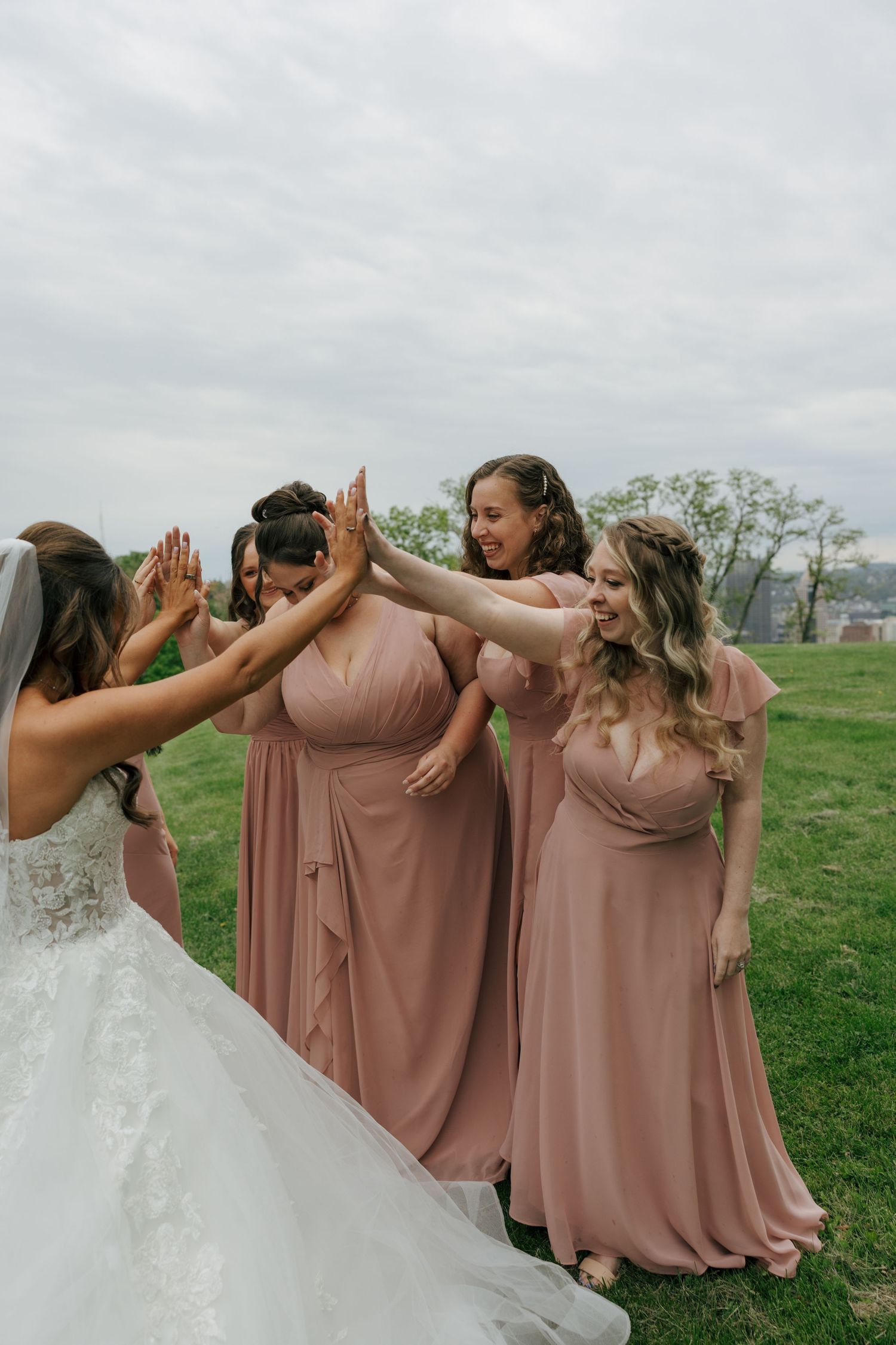 Bride and bridesmaids in dusty rose gowns celebrate with a group high-five on a green lawn.
