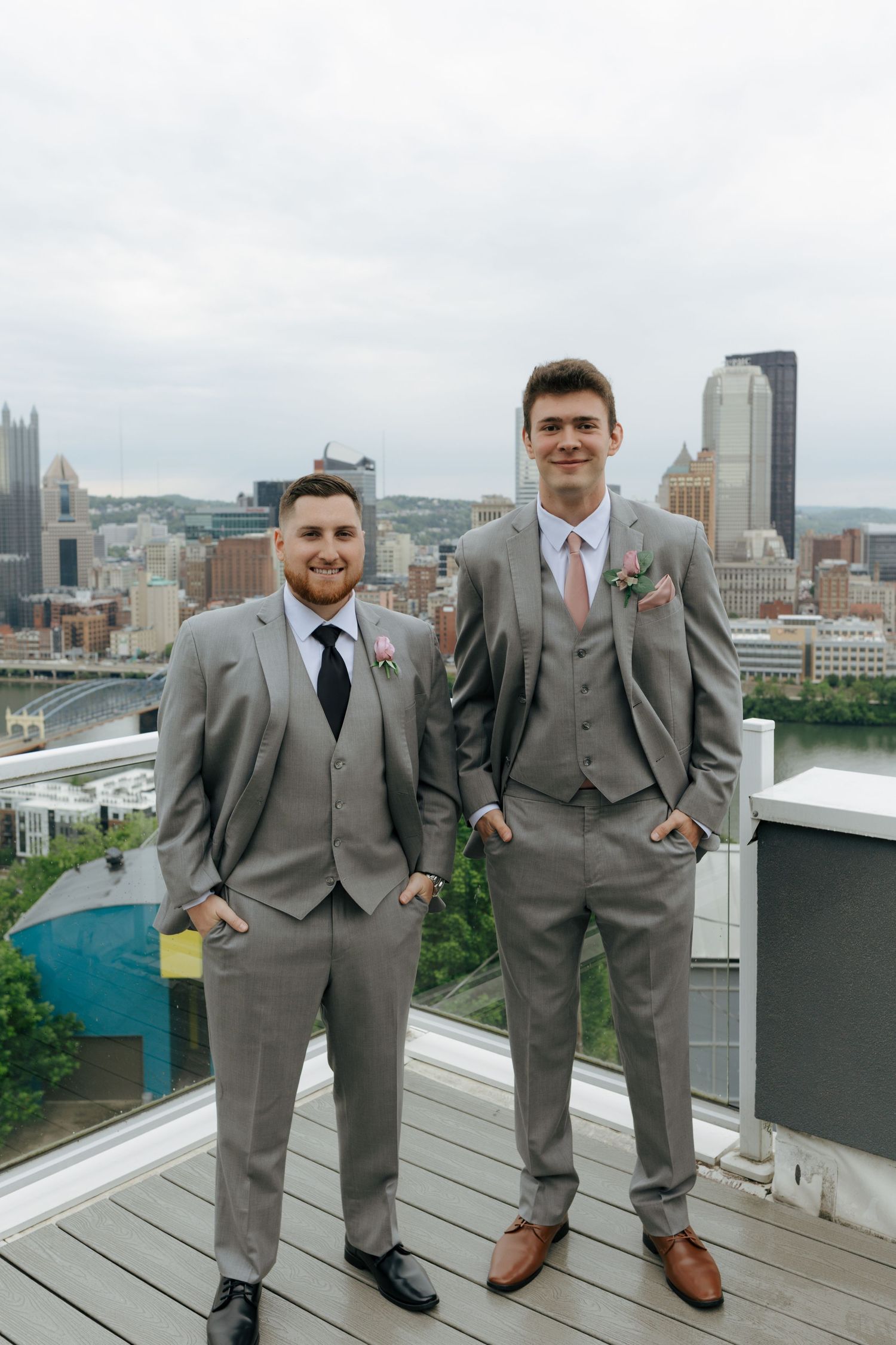 Two groomsmen in gray suits with boutonnieres pose on a rooftop with Pittsburgh skyline behind them.