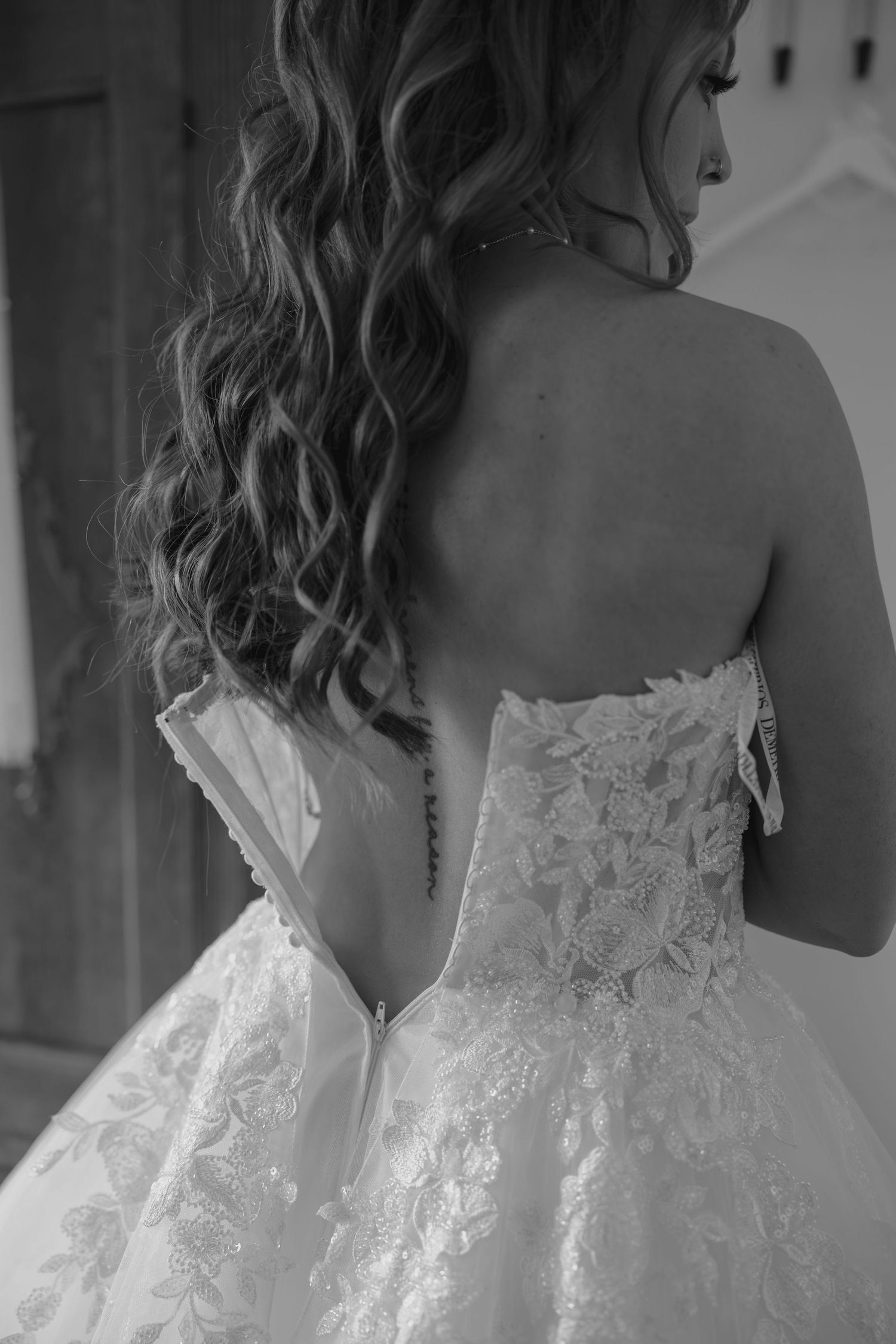 Black and white photo of a bride in a strapless lace wedding gown, showing her back with curly hair and a small tattoo.