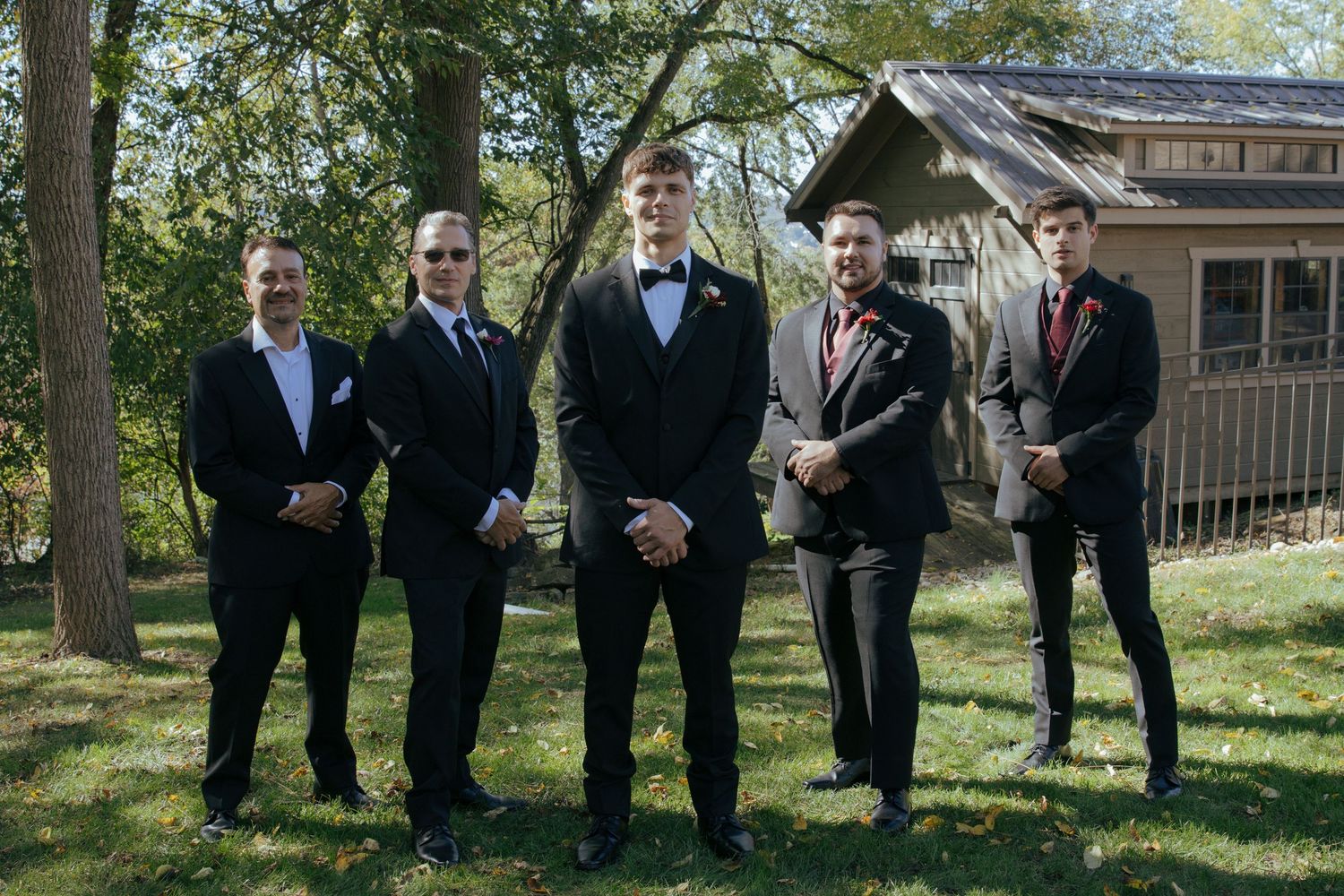 Five groomsmen in black tuxedos pose outdoors on green grass near a rustic wooden building.