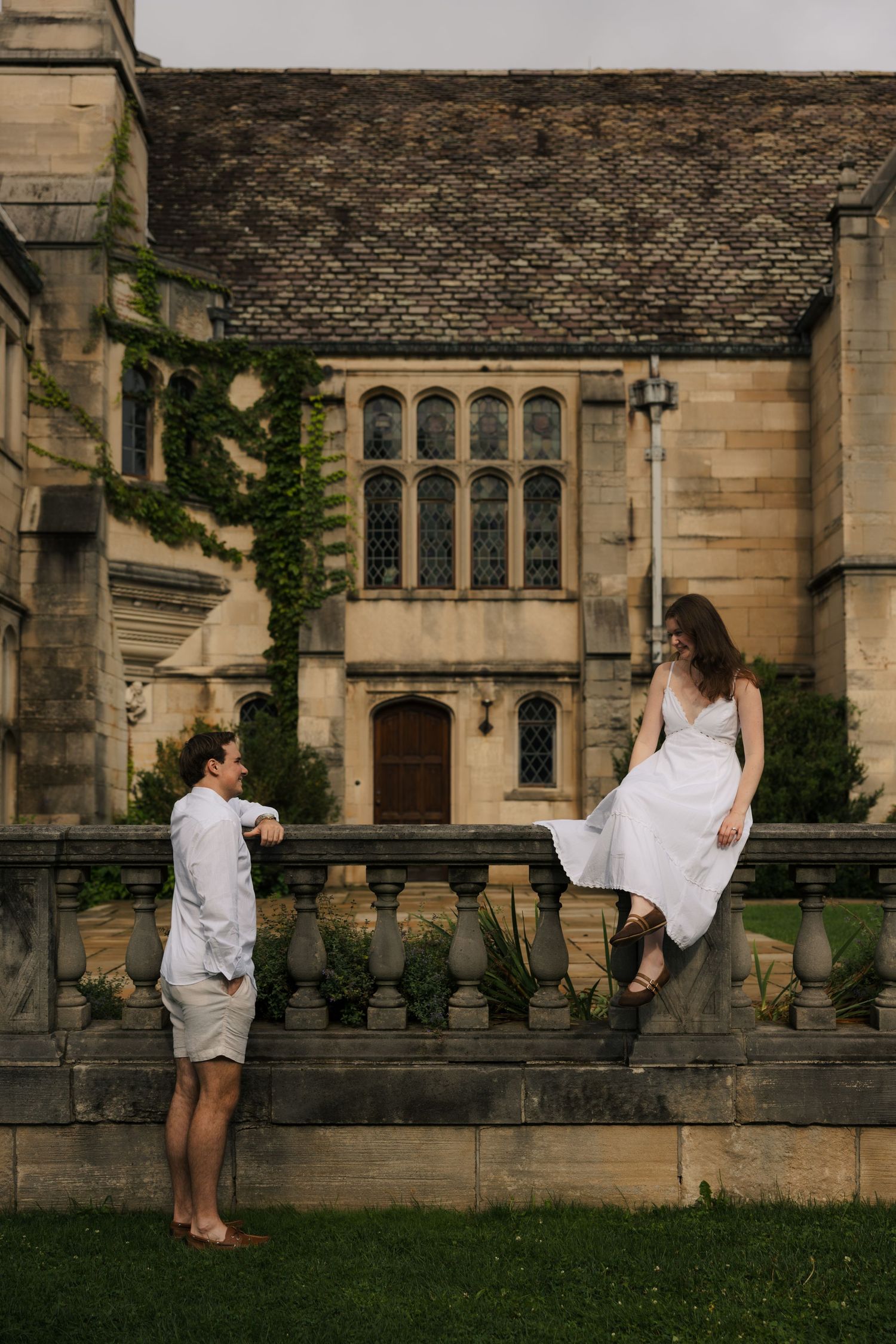 Young couple in matching white outfits interacting near ornate stone balustrade of ivy-covered historic building.