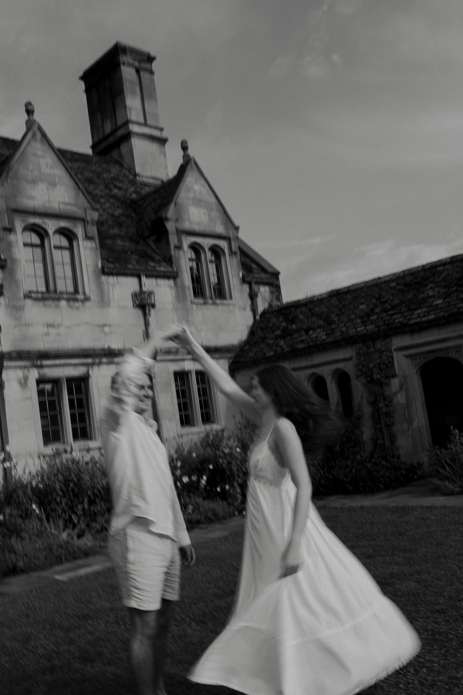 Blurred black and white photo of a couple dancing joyfully on the lawn of a grand stone manor house.