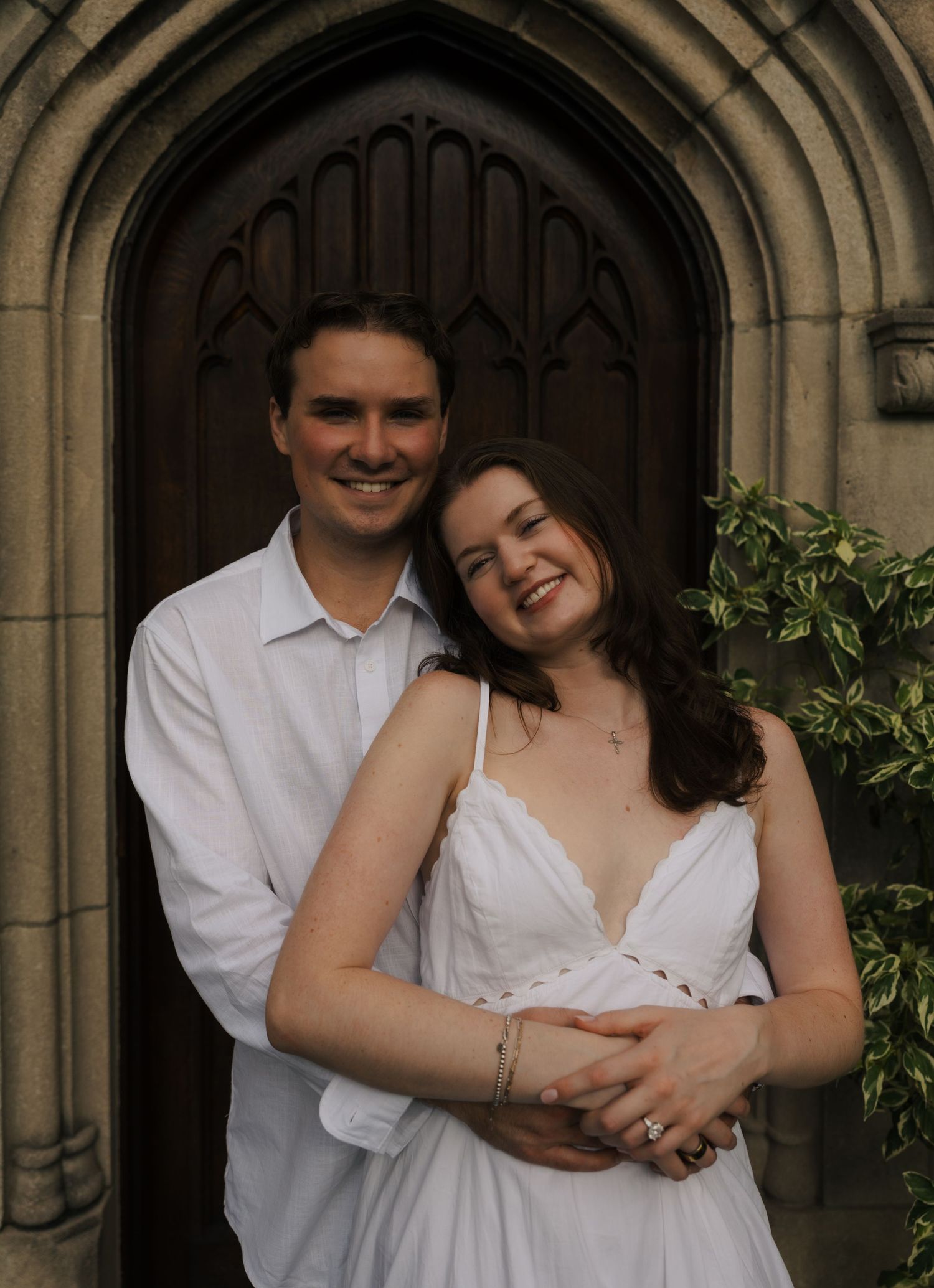 Smiling couple in white outfits embracing before ornate gothic arched wooden church door.