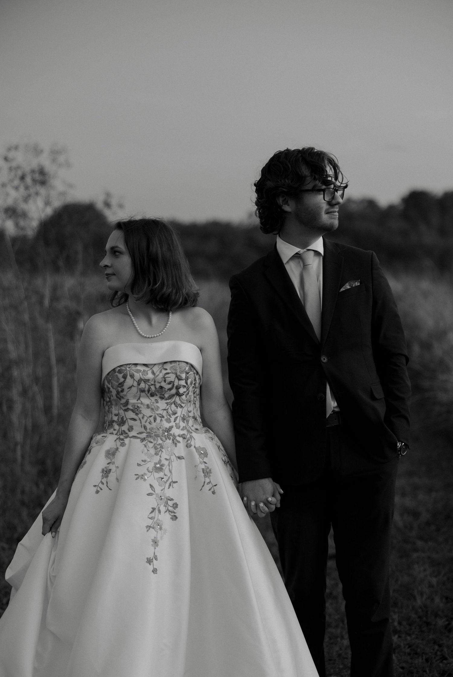 Black and white photo of bride and groom holding hands in a field at dusk, wearing elegant wedding attire.