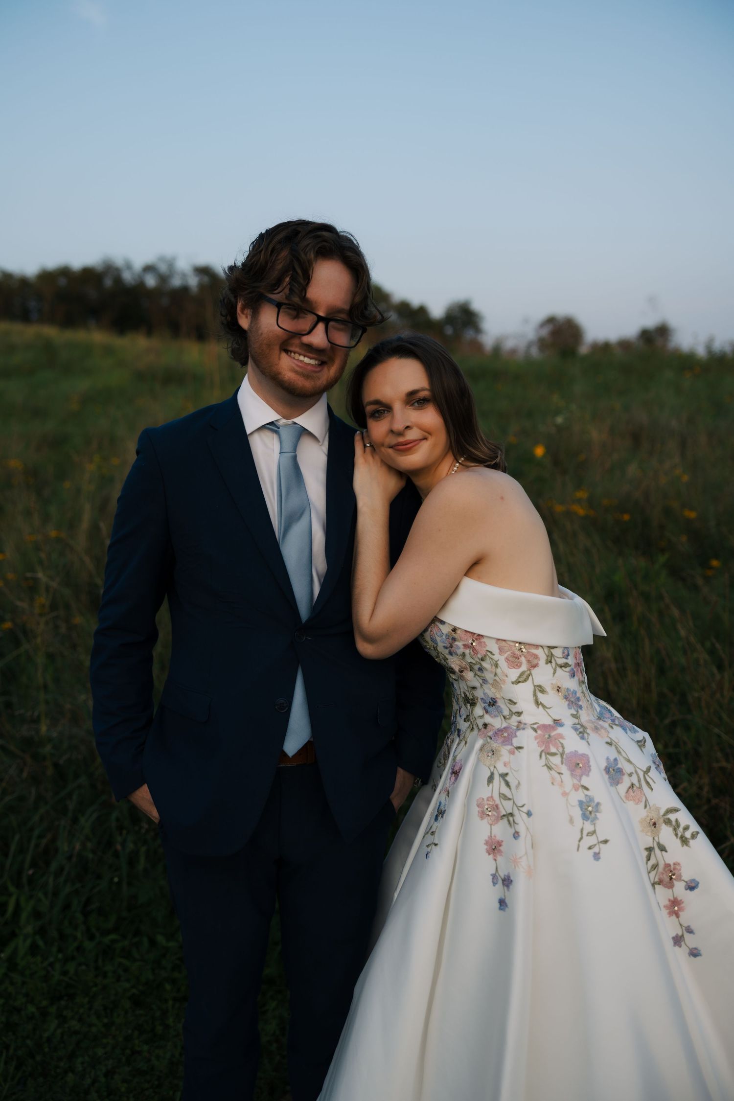 Wedding couple posing in a green field at sunset, bride in floral off-shoulder gown, groom in navy suit.