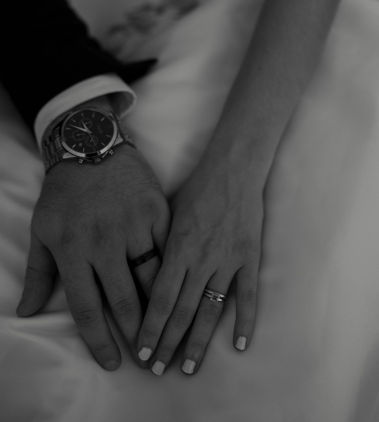 Black and white close-up of bride and groom\u2019s hands, showcasing wedding ring and groom\u2019s classic dress watch.