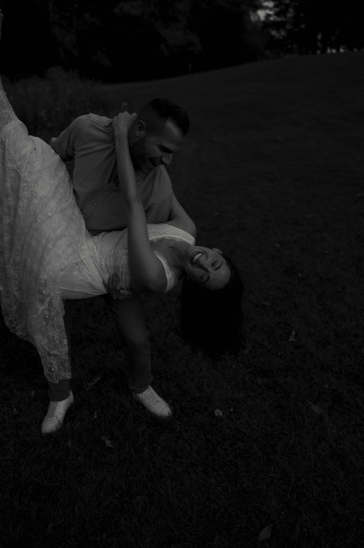 Black and white photo of a man dipping a laughing woman in a white dress on a grass lawn at night.