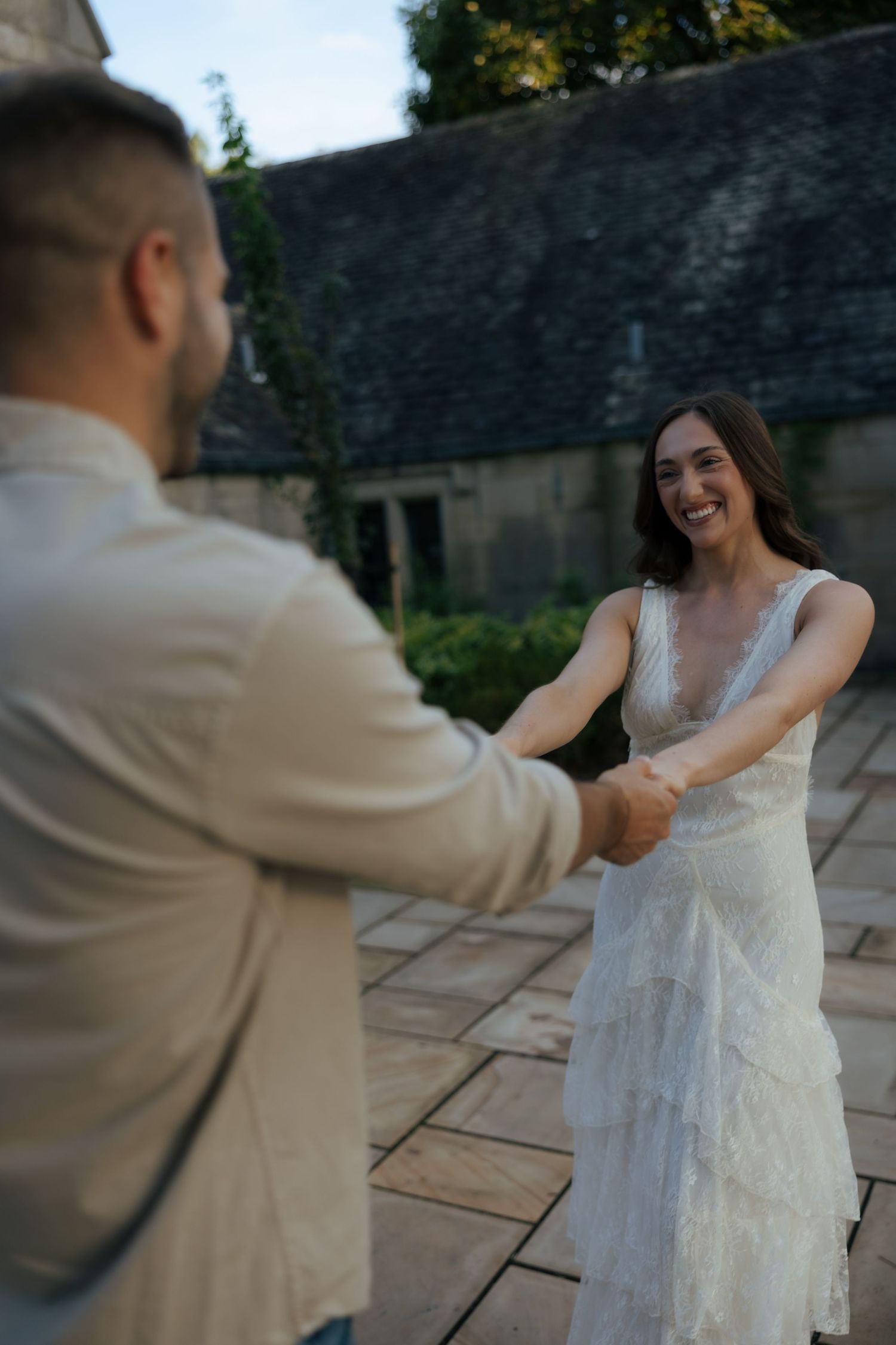 Smiling bride in white lace dress holds hands with groom in beige shirt on stone patio at outdoor wedding venue.