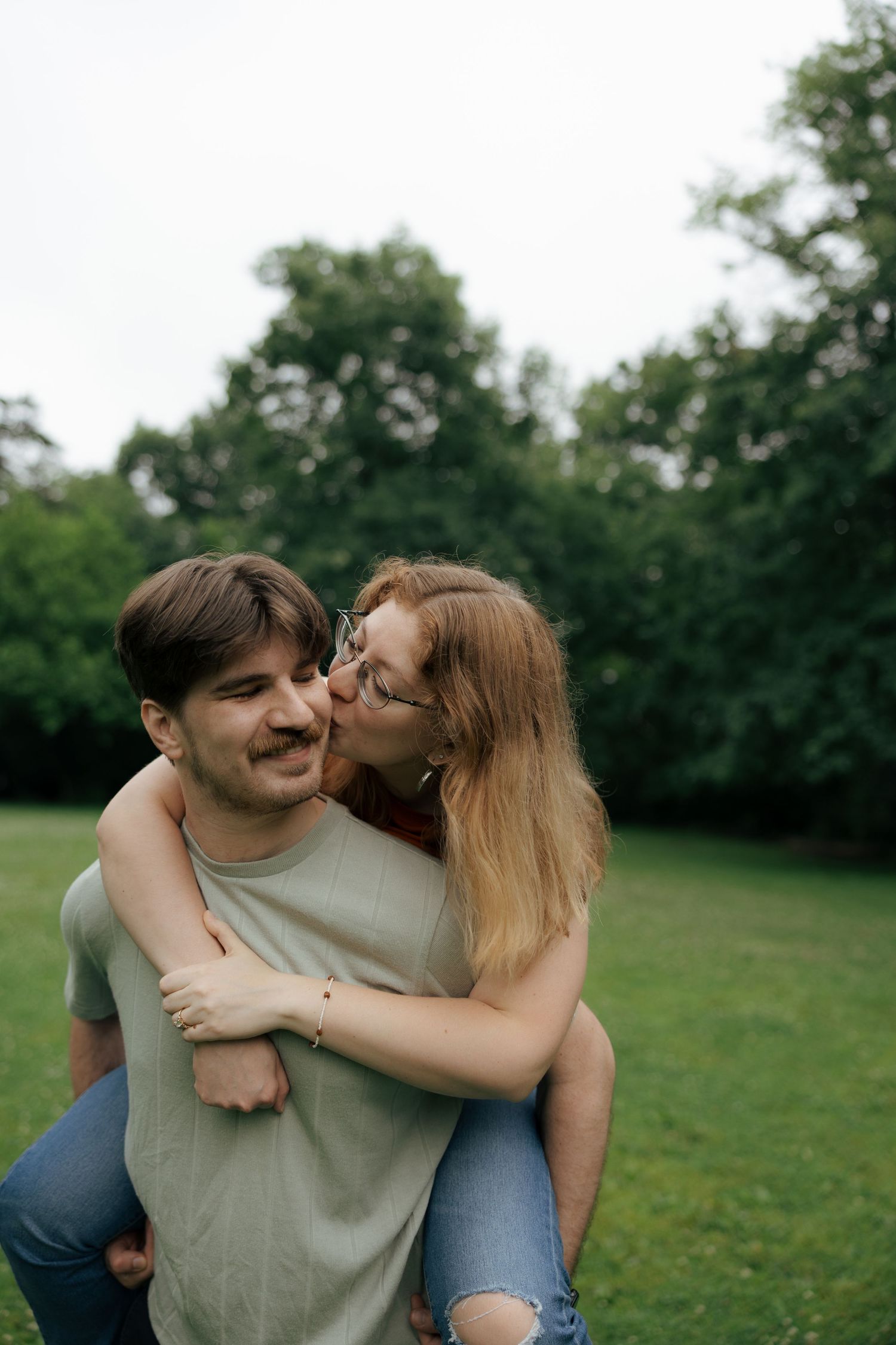 A woman kisses a man on the cheek as he gives her a piggyback ride in a green park.