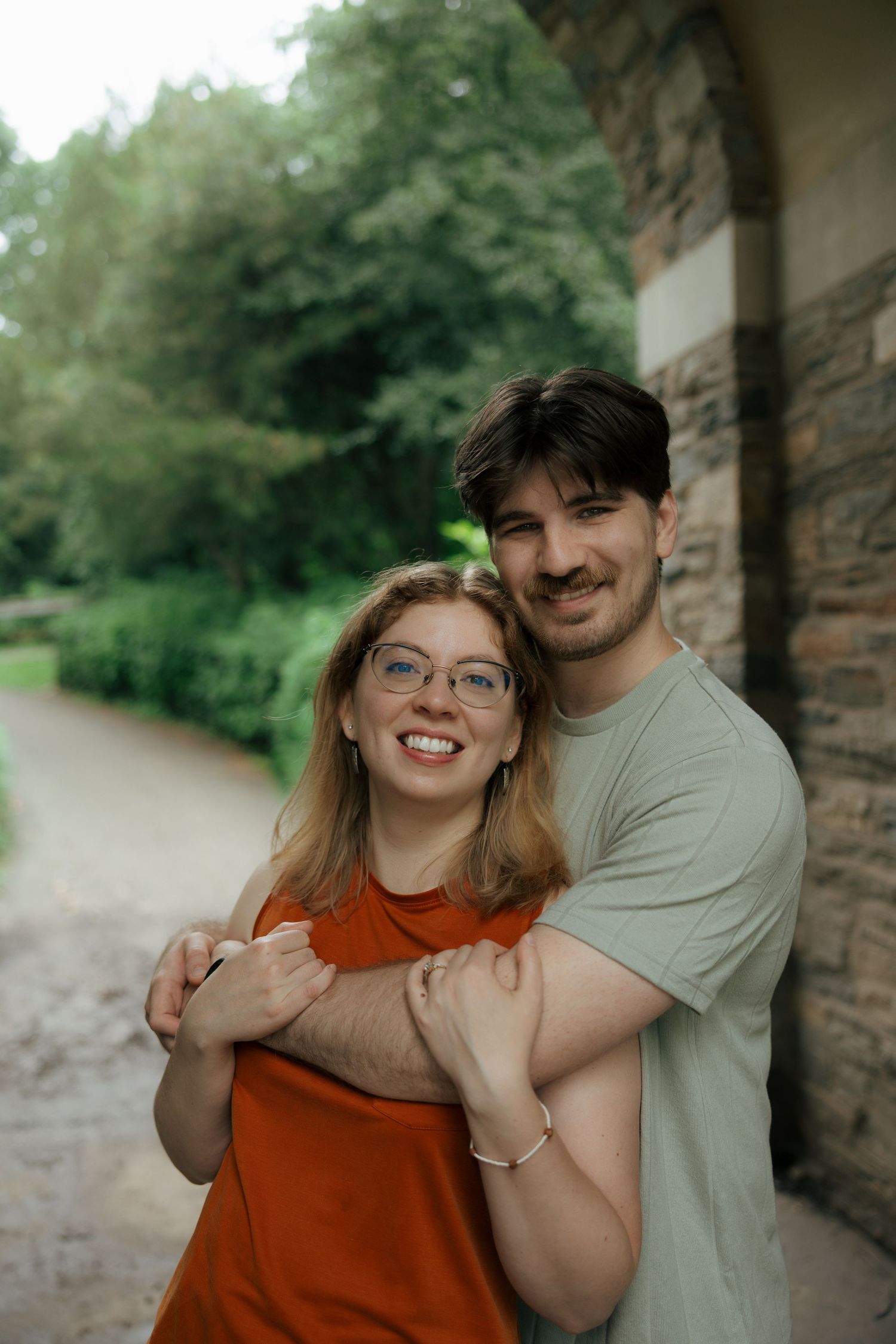 Smiling couple embraces near stone wall surrounded by lush green trees.