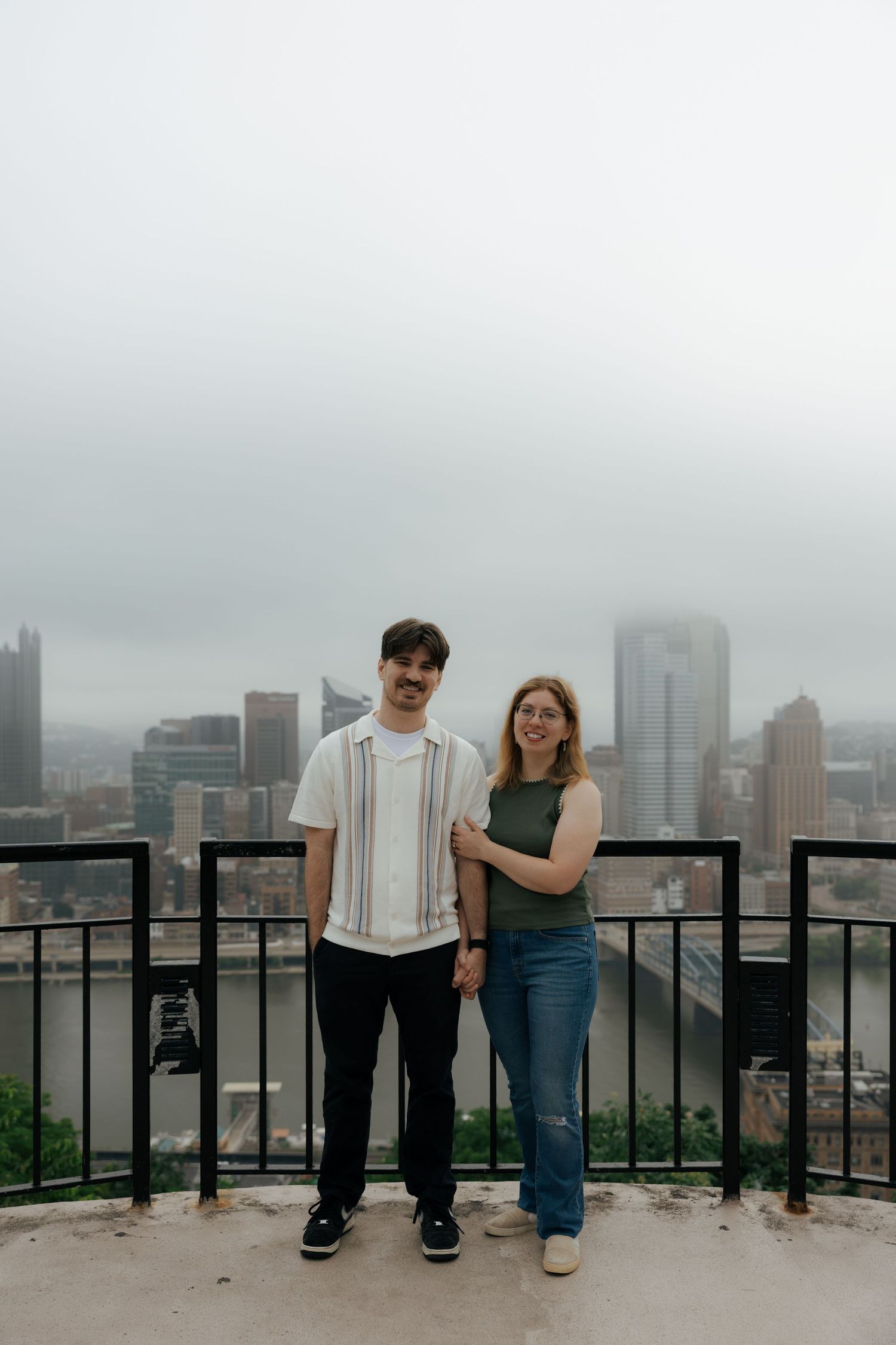 Couple posing together on a scenic overlook with a misty Pittsburgh skyline in the background.