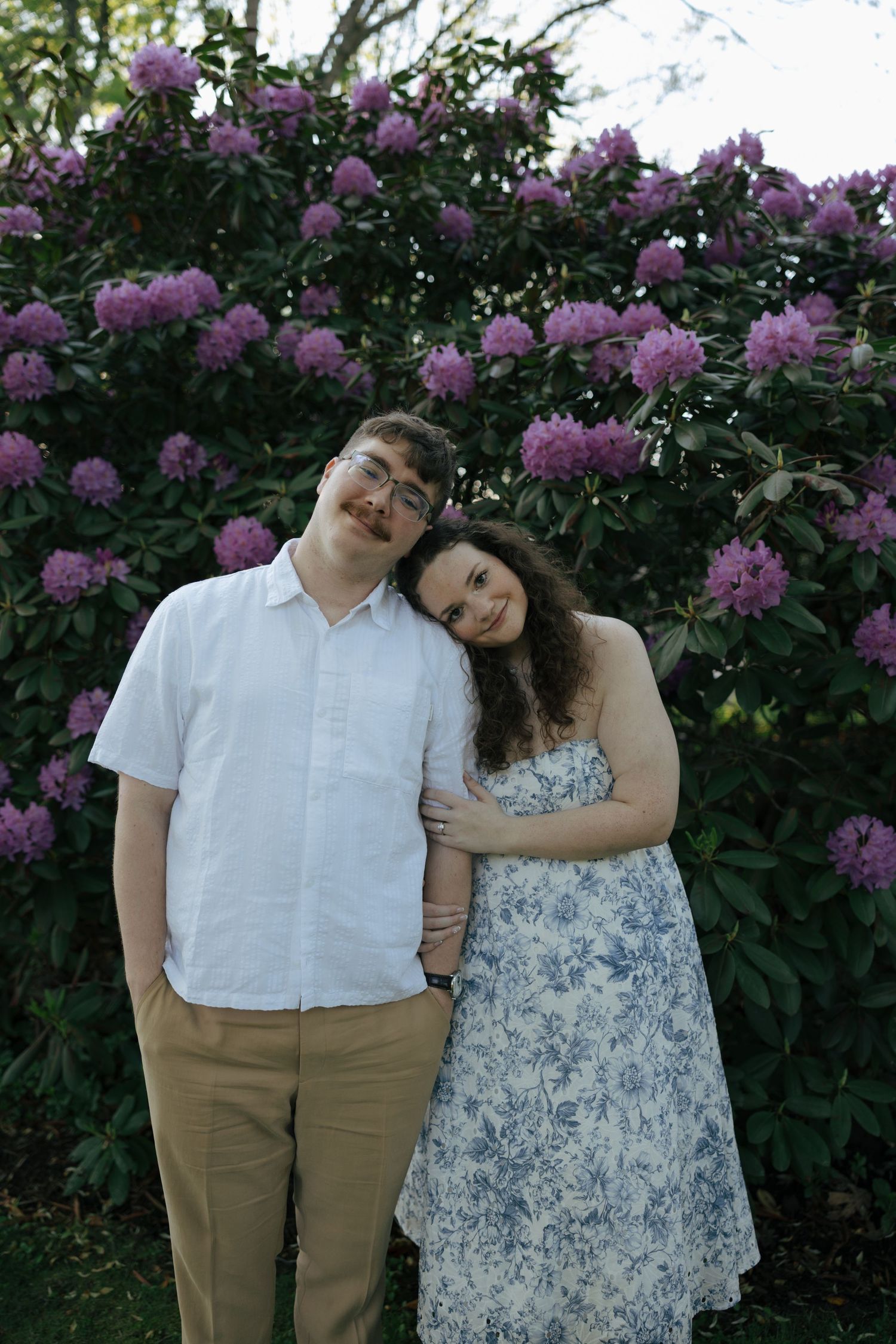 Engaged couple smiling together in front of blooming purple rhododendron bushes, woman in floral dress.