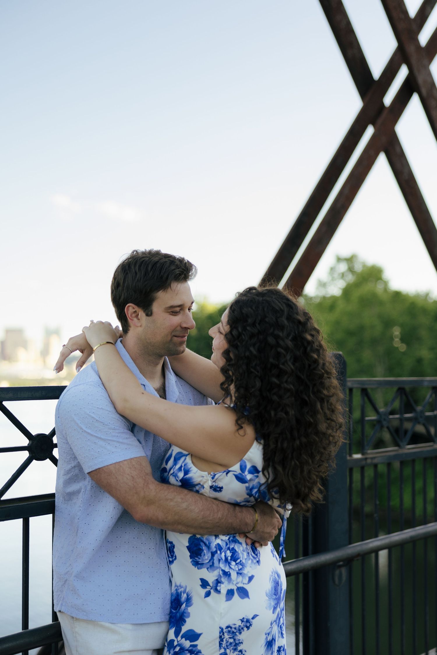 Couple embracing and gazing at each other on a bridge with city skyline behind them.