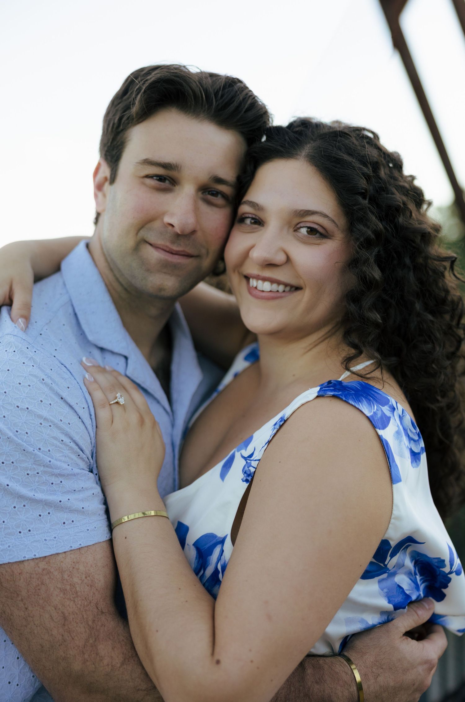 Smiling engaged couple embracing closely, woman wearing diamond ring and blue floral dress outdoors.